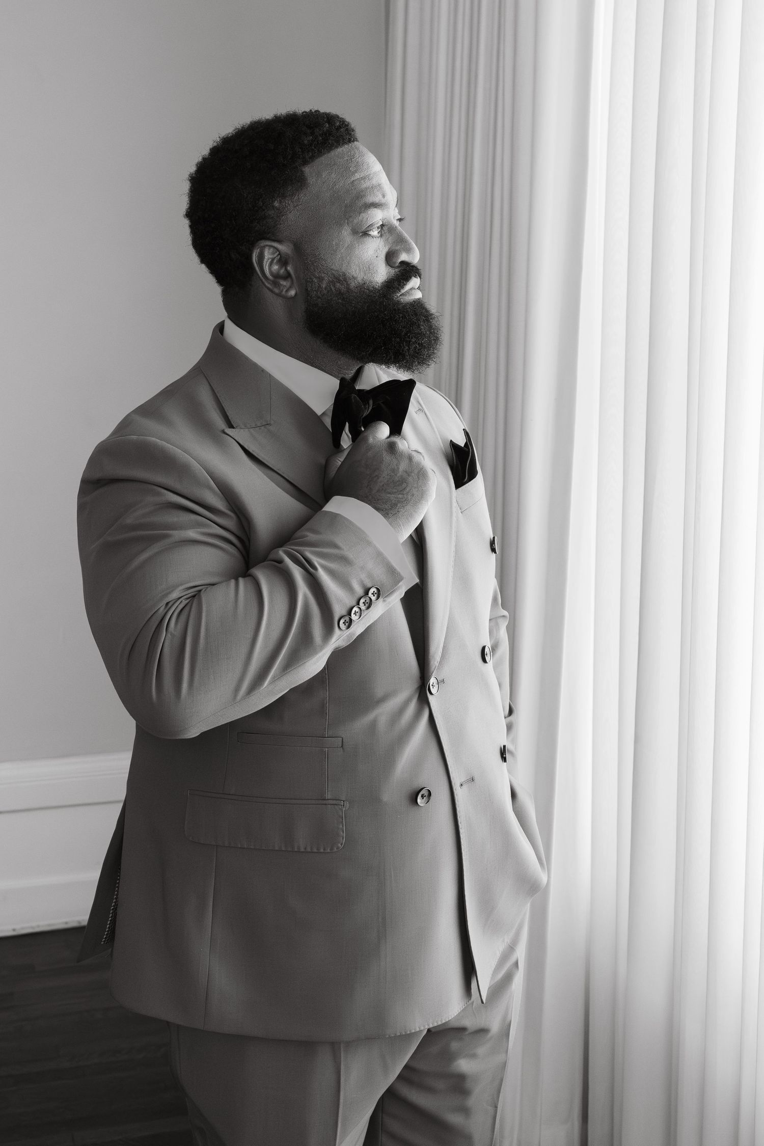 Groom in double-breasted suit adjusting bow tie by window in black and white portrait photo.