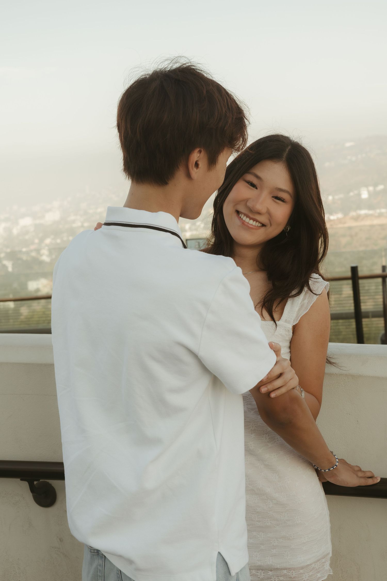 Smiling Asian couple in white outfits sharing a romantic moment on a scenic overlook balcony.