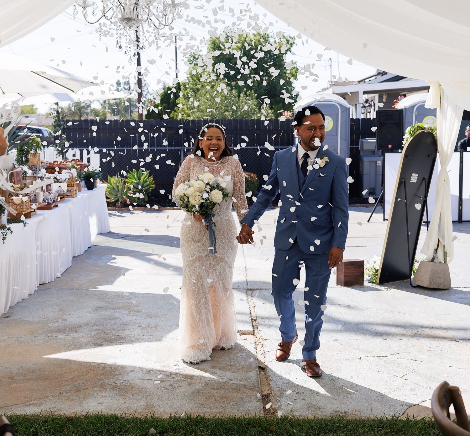 Newlyweds hold hands and smile as guests toss petals during their outdoor wedding exit, with the bride carrying a white bouquet.