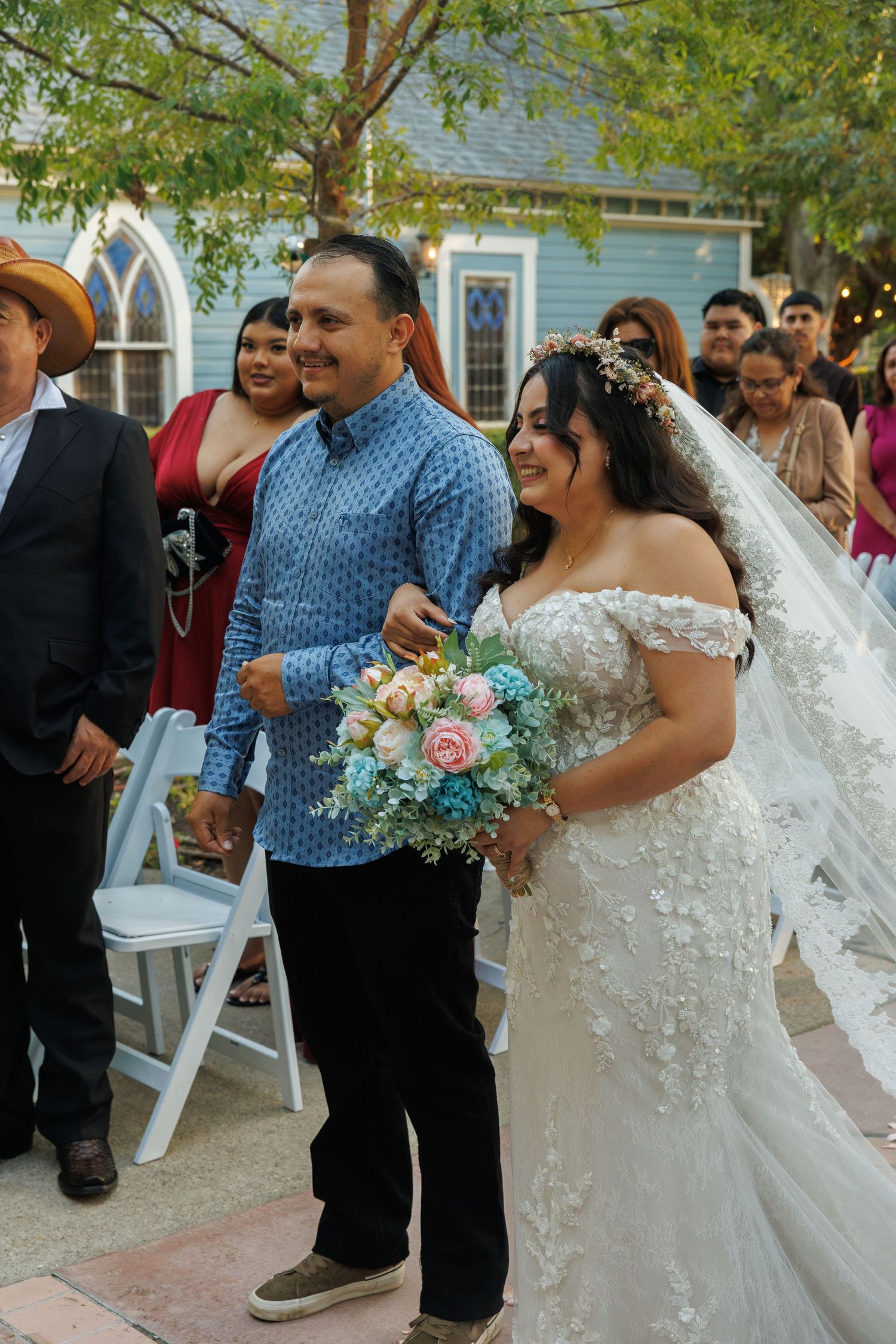 Bride in white lace gown holding floral bouquet walks down aisle with escort at outdoor wedding ceremony.