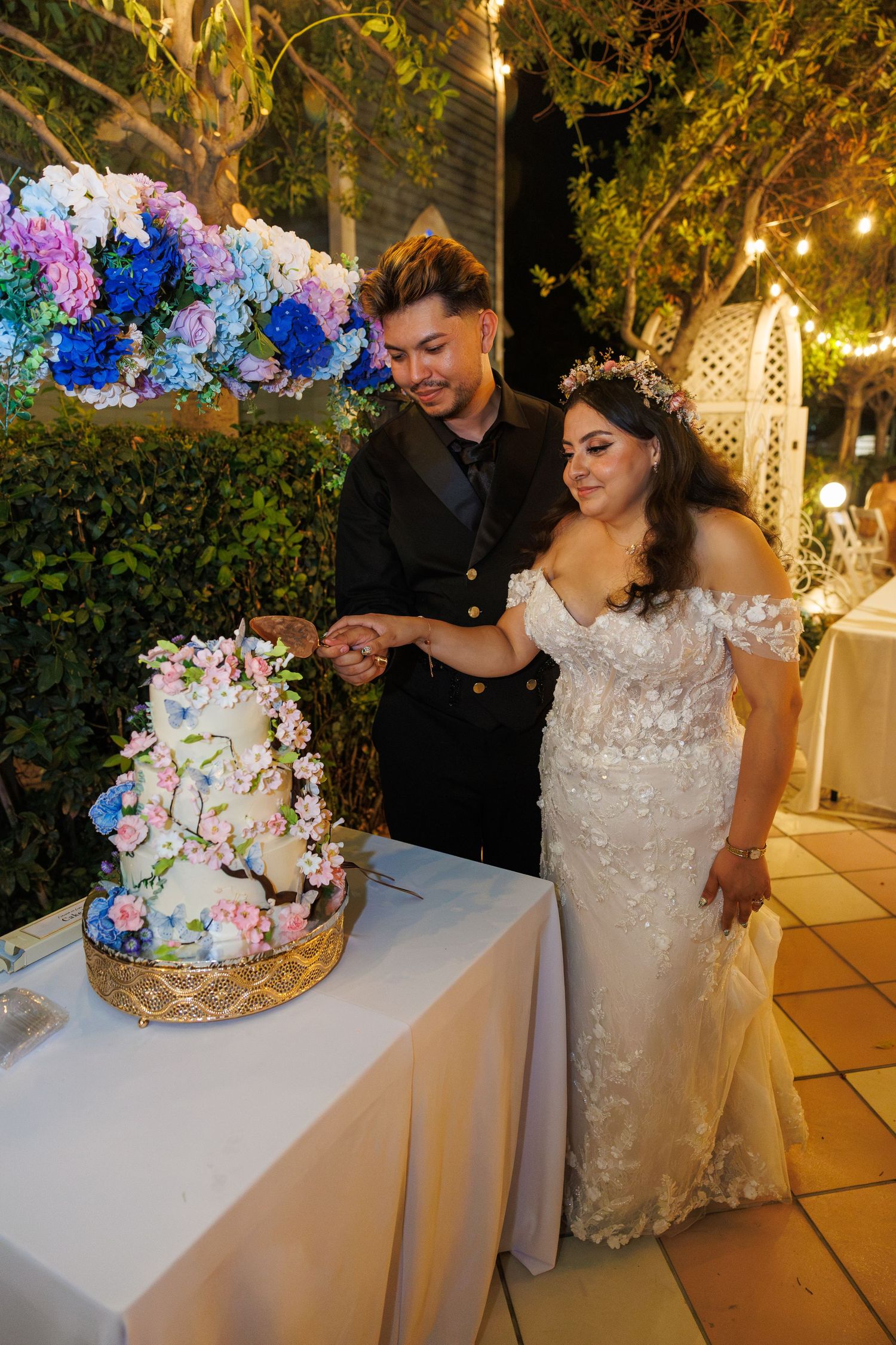 Bride and groom cut a floral-decorated wedding cake outdoors under warm string lights at night.