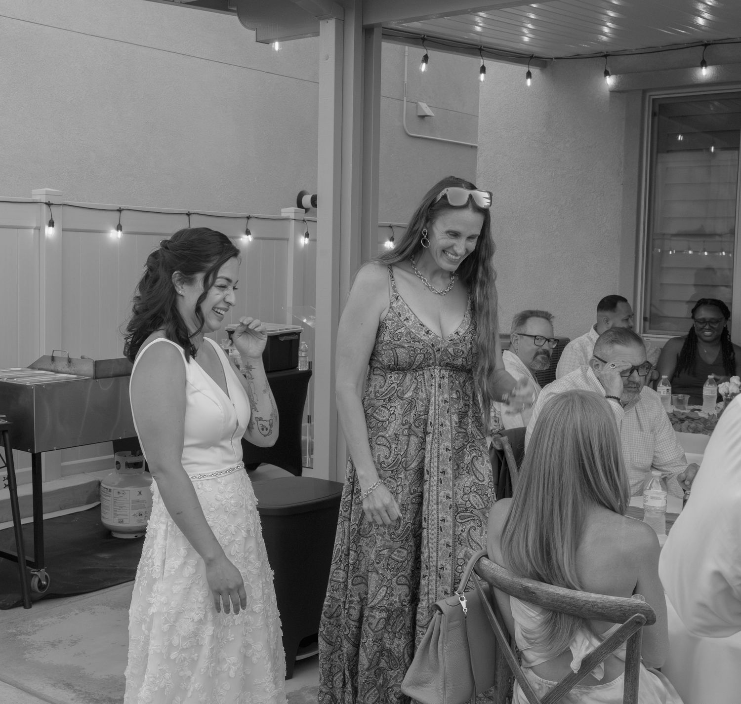 Black and white photo of two women in elegant dresses socializing at an indoor event with string lights.