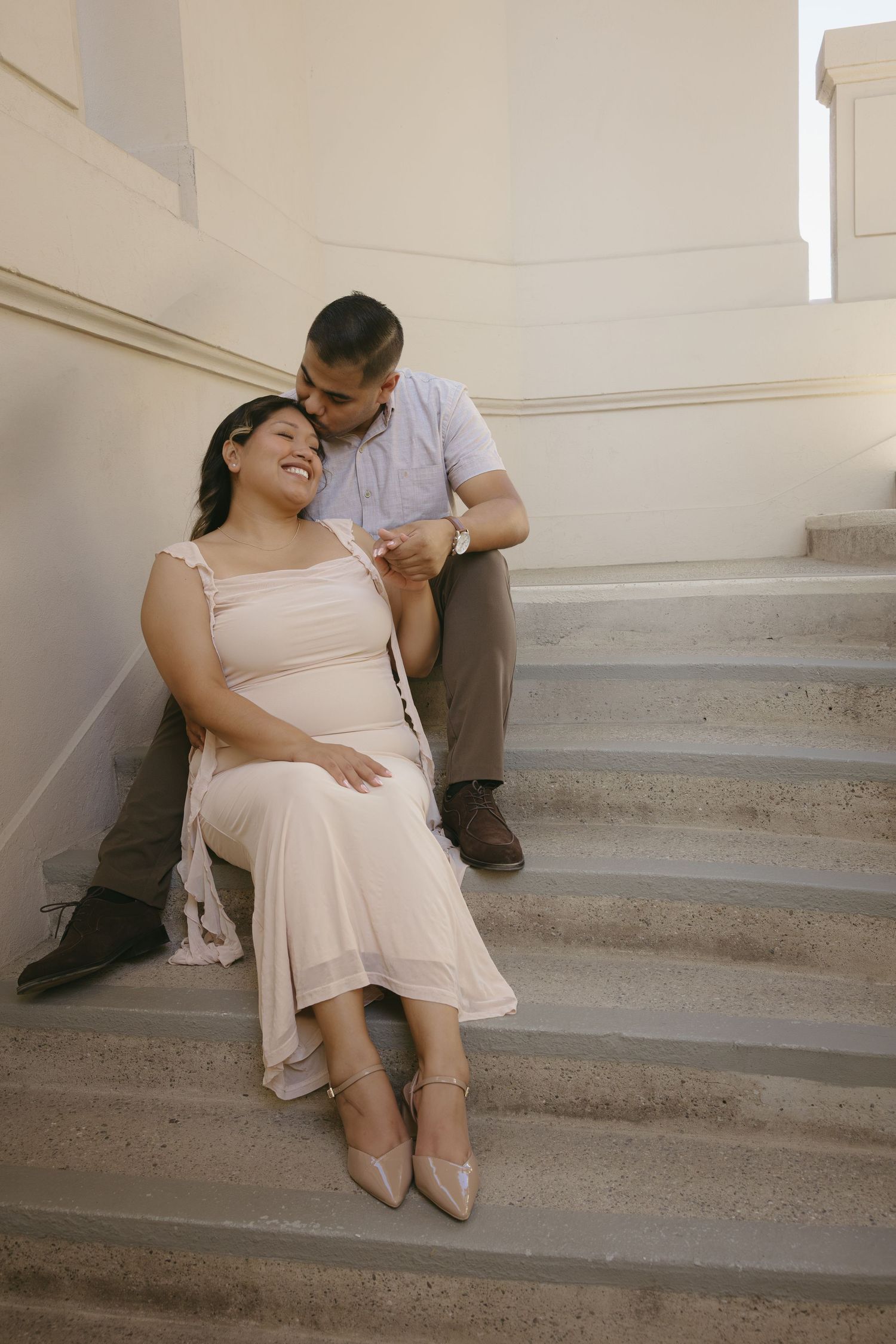Couple sharing tender moment on stone steps, pregnant woman in blush dress leaning against partner in soft sunlight.