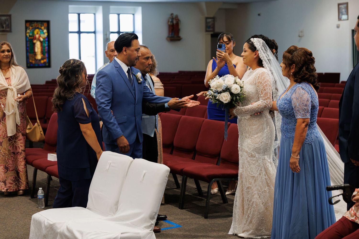 A bride in a white gown and veil holds a bouquet while guests in formal attire gather in a church with red pews during a wedding ceremony.