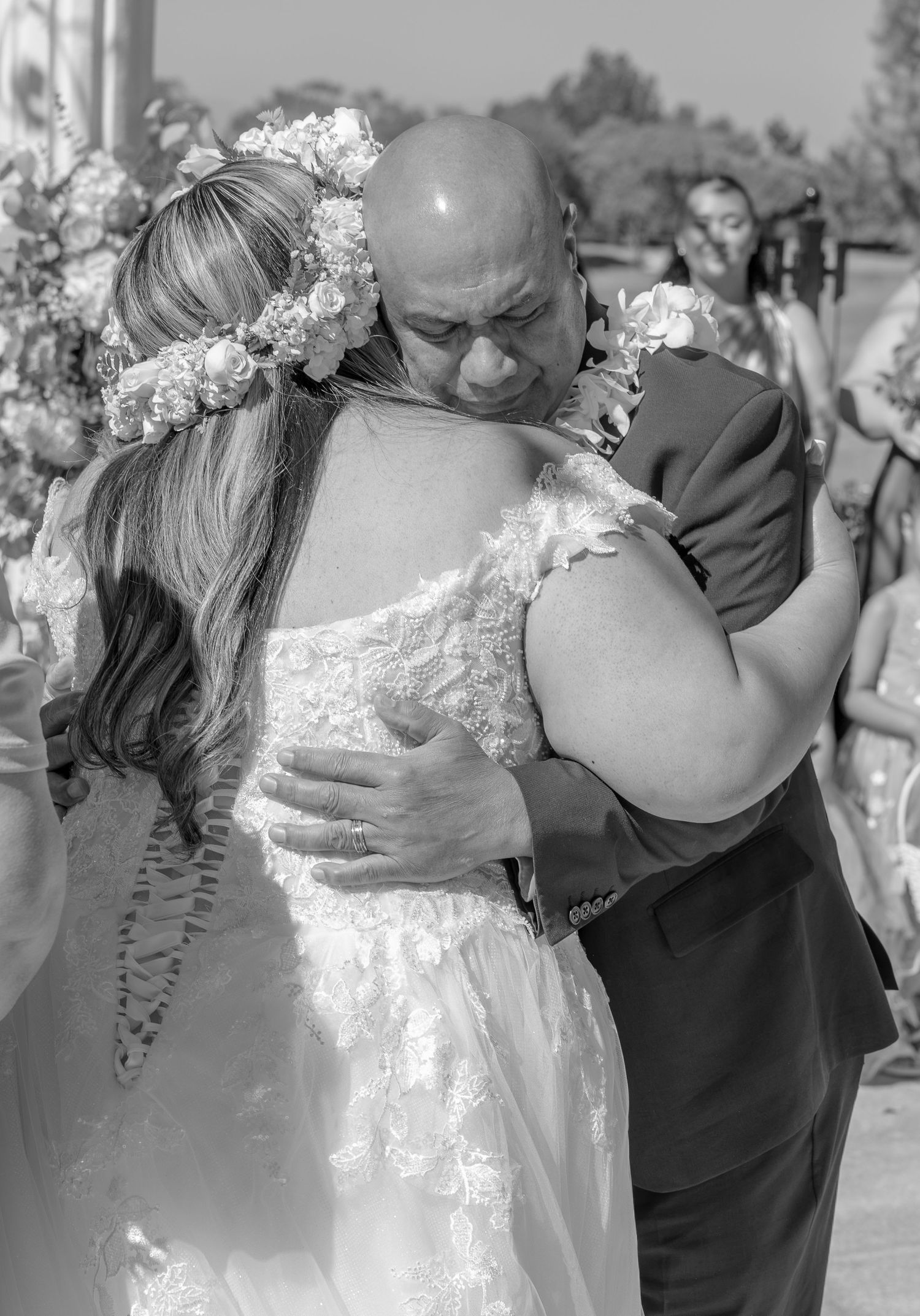 Emotional black and white photo of bride in floral crown embracing bald man at outdoor wedding.