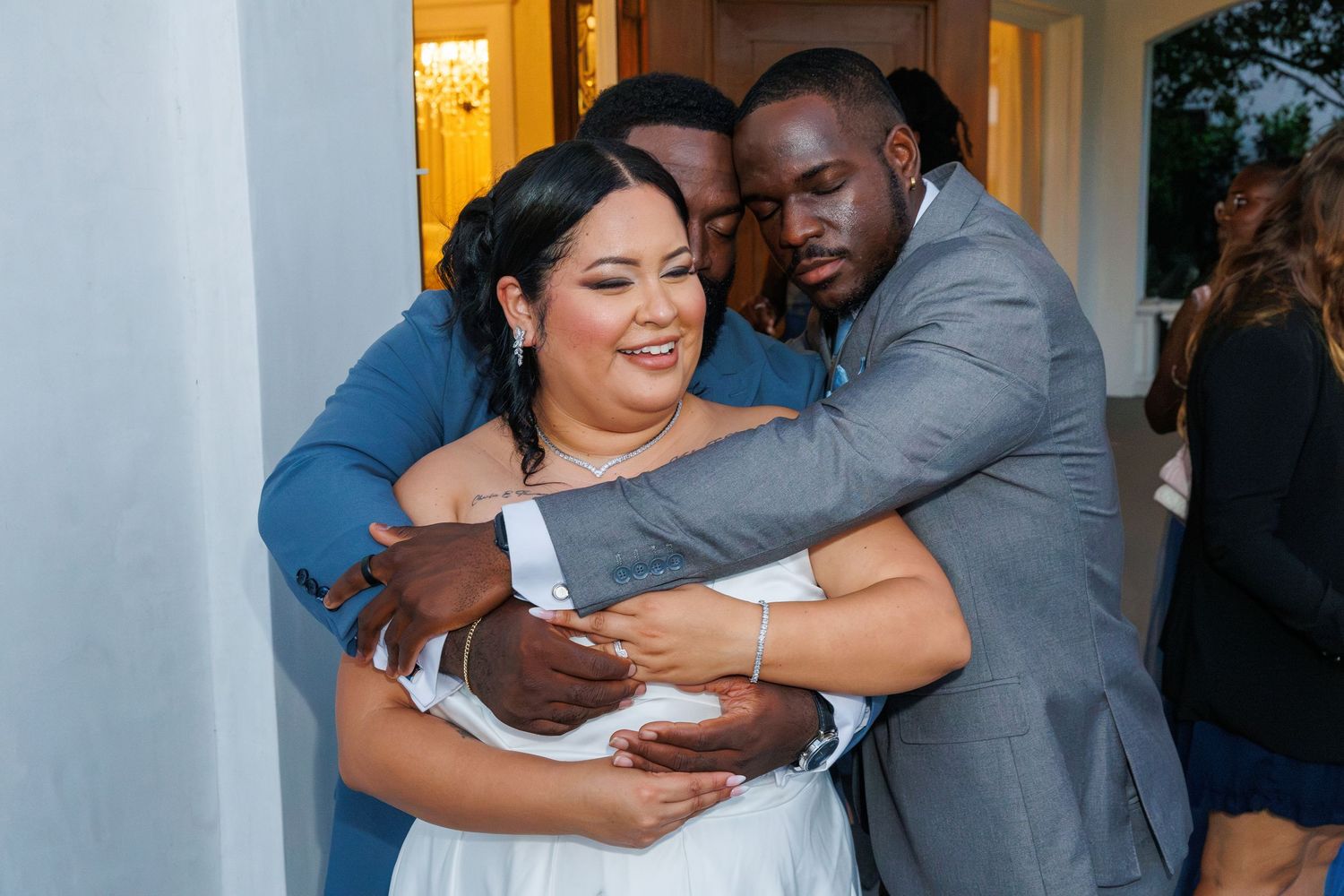 Bride and groom embrace warmly with a guest holding a baby at wedding reception.