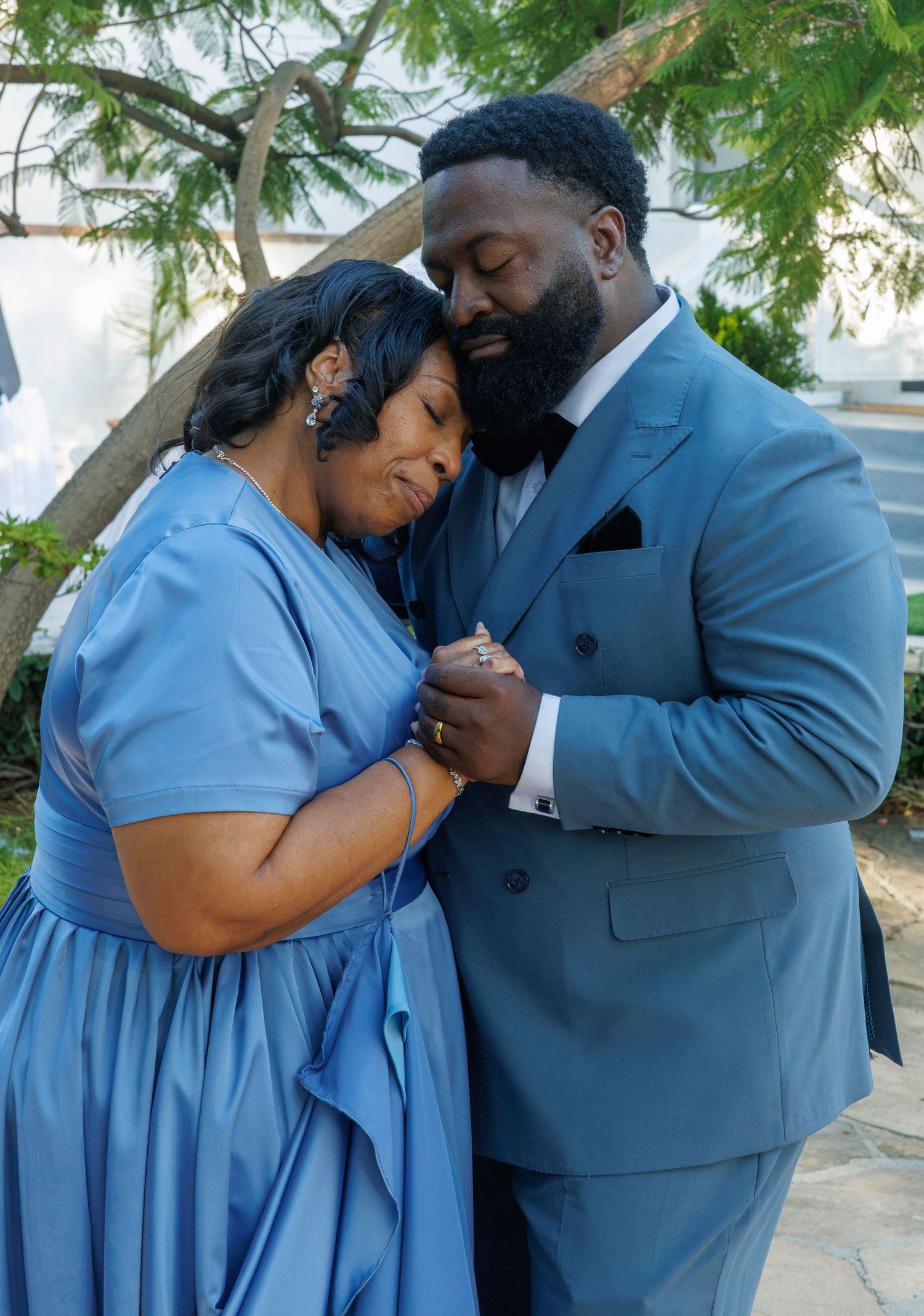 Emotional mother in blue dress leans on son in blue suit, holding hands tenderly at an outdoor wedding.