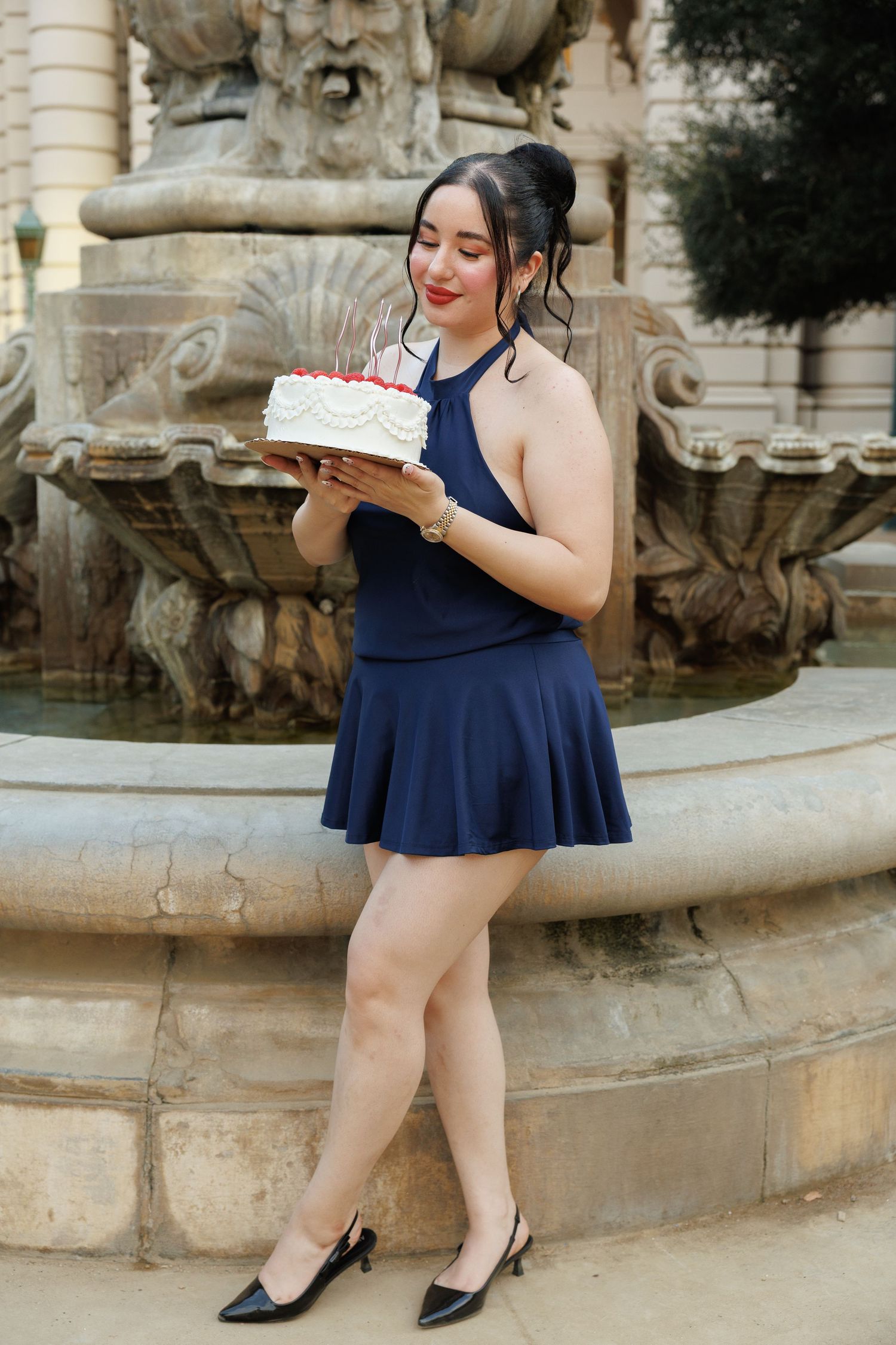 Young woman in navy blue dress holding a birthday cake near an ornate stone fountain outdoors.