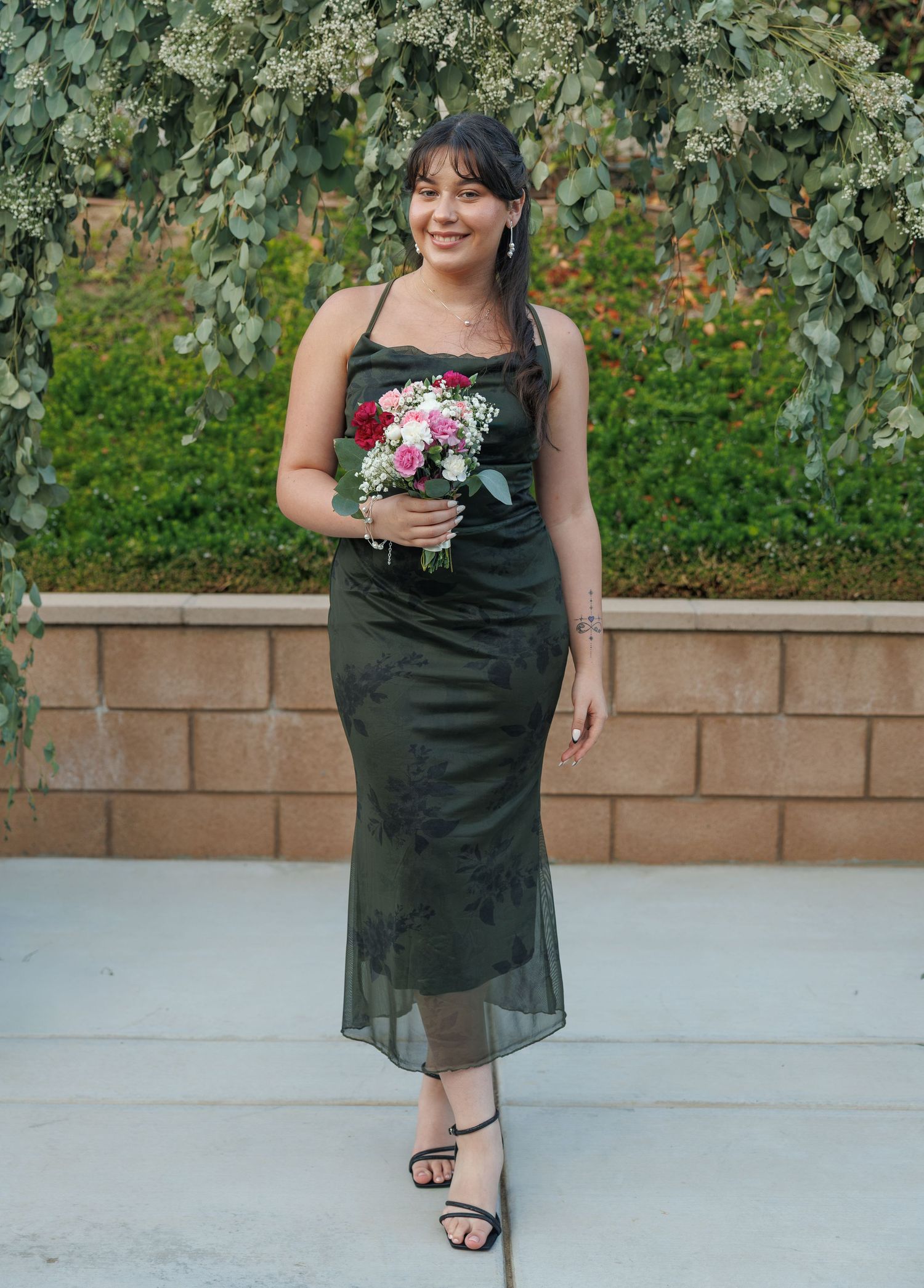 Woman in dark green midi dress holding a floral bouquet, posing outdoors with lush greenery backdrop.