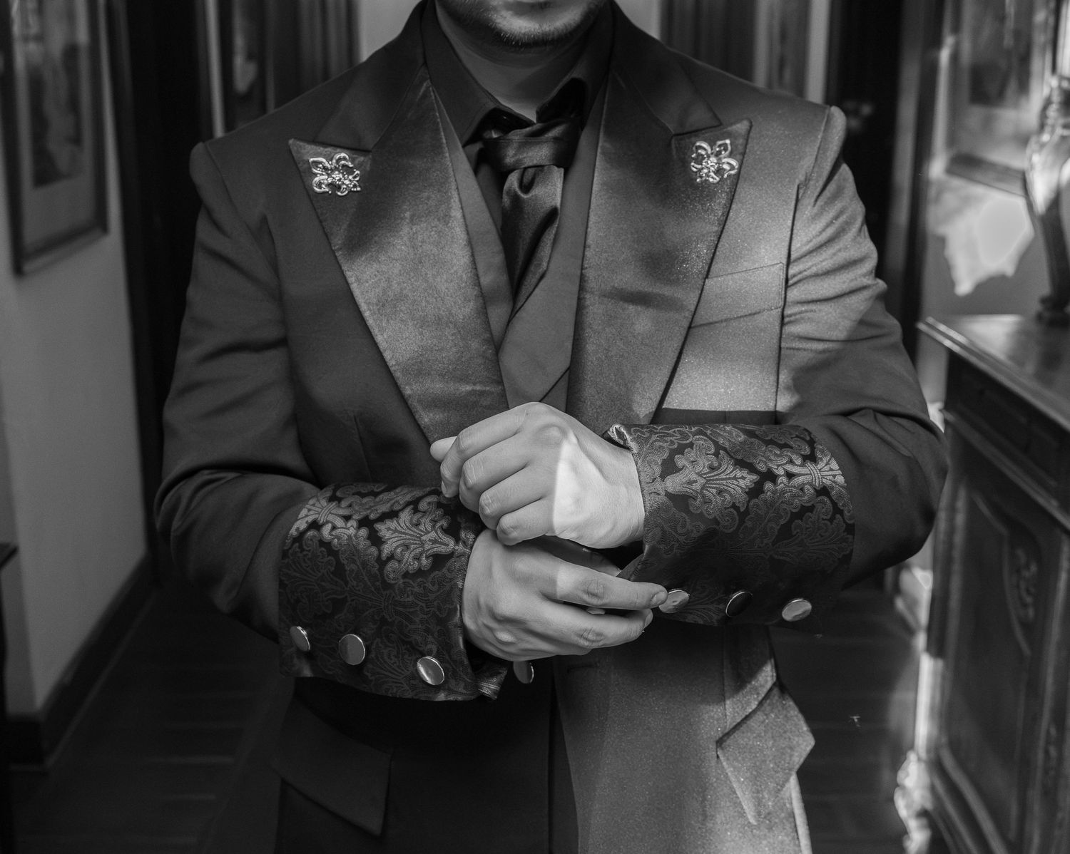 Man in ornate suit with decorative lapel pins adjusting cufflinks, black and white formal fashion photography.