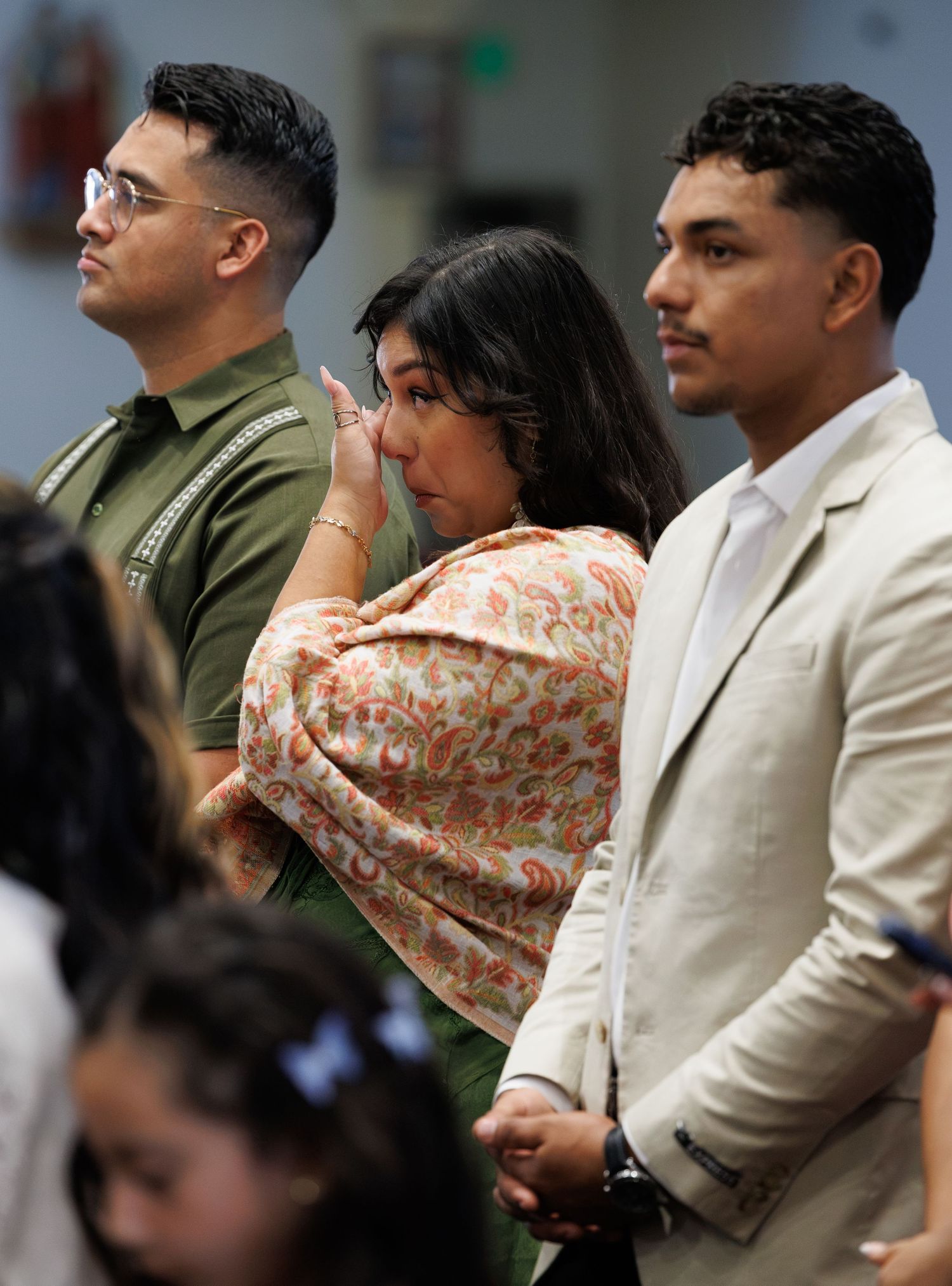 A woman in a floral shawl wipes her eye while standing between two men in a crowd, appearing emotional during a gathering.