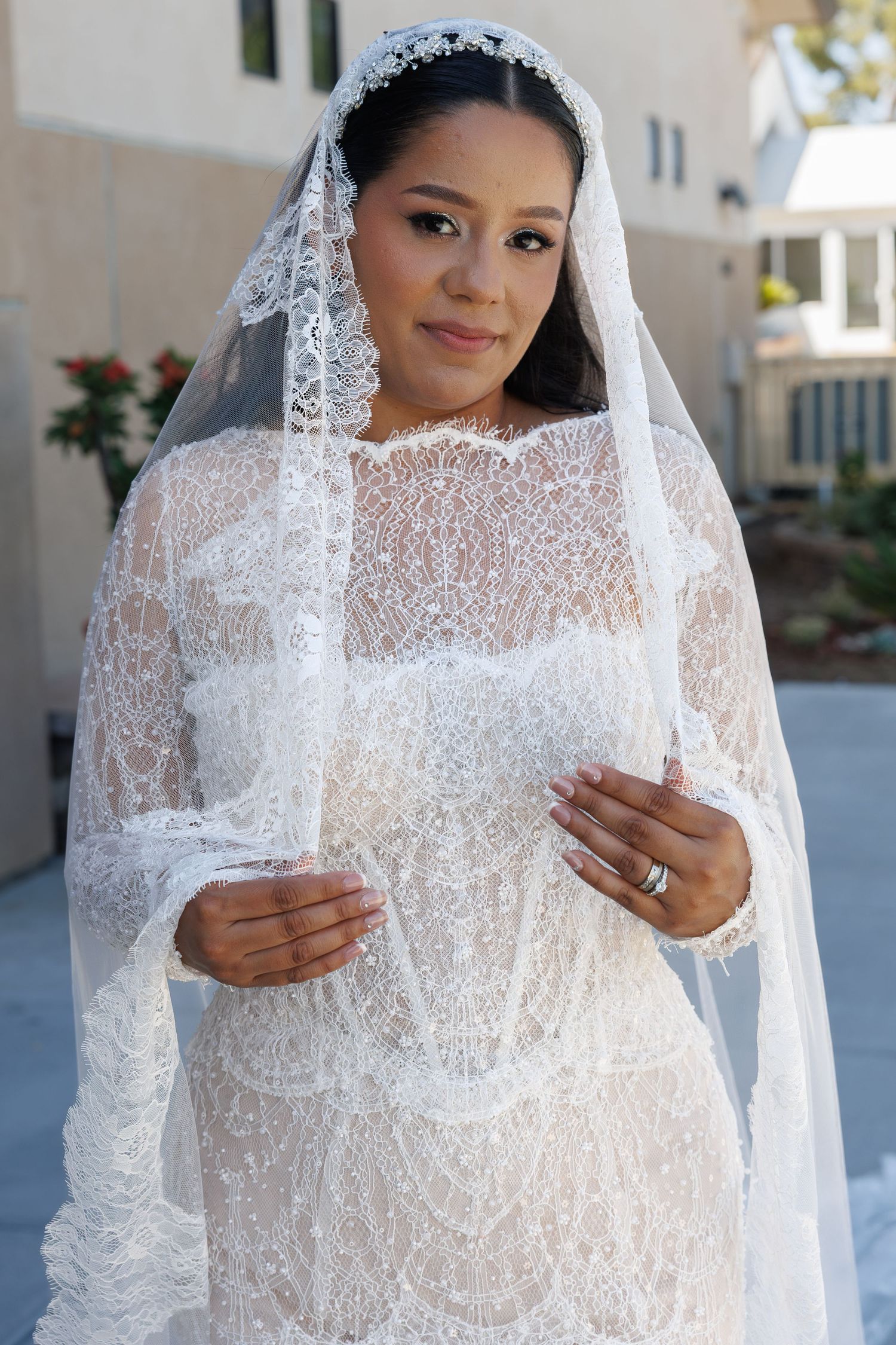 Smiling bride in an ivory lace wedding gown and matching lace-trimmed cathedral veil poses outdoors, hands gently clasped at her chest.