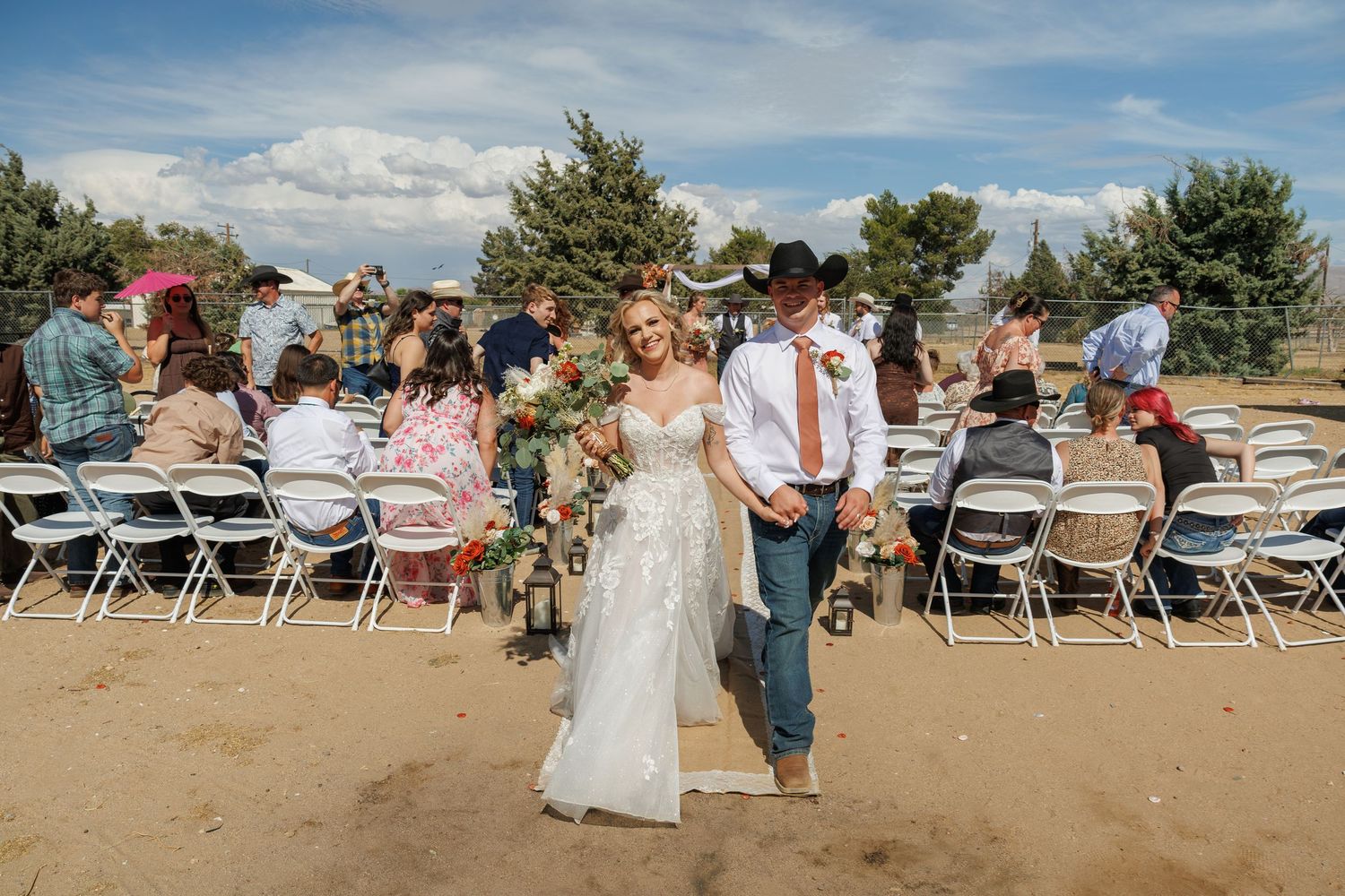 Happy newlywed couple in western attire pose together after outdoor rustic wedding ceremony with white chairs.