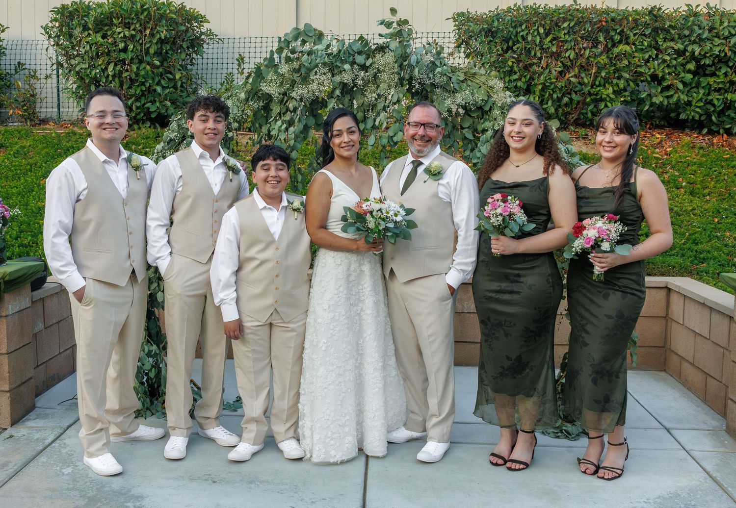 Wedding party poses outdoors, bride in white gown, groom in suit, flanked by family in coordinated beige and green attire.