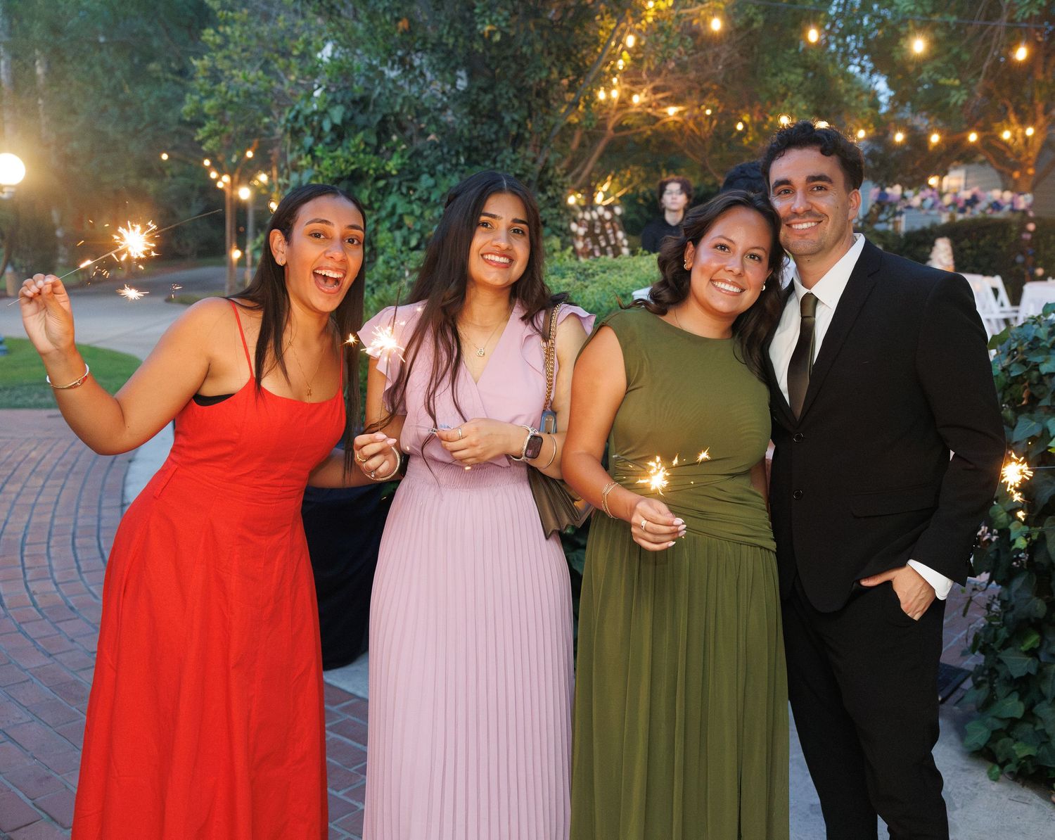 Four friends in formal attire smile holding sparklers at an outdoor evening wedding celebration.