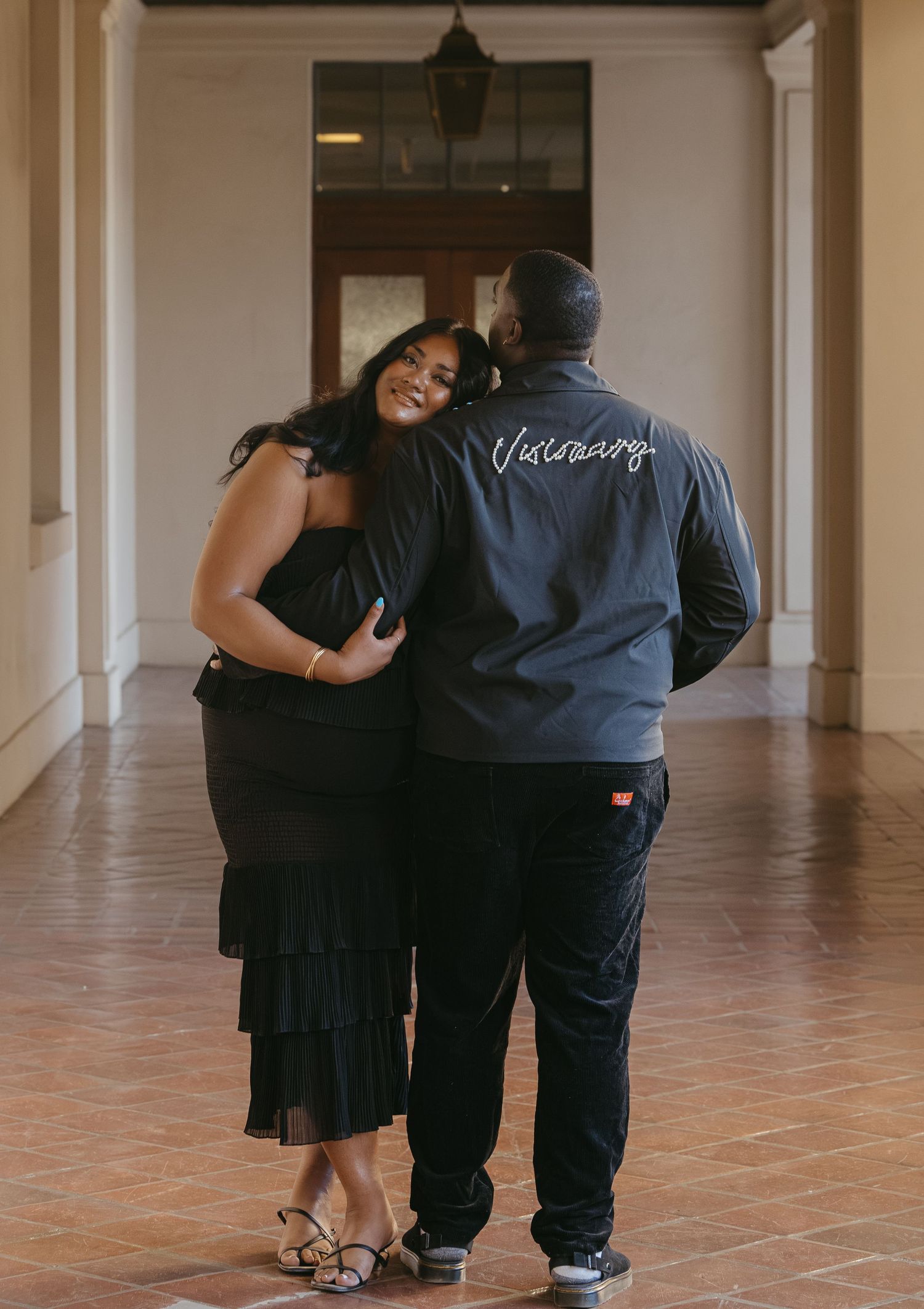 Couple embracing in elegant hallway, woman in black ruffled dress, man in gray jacket with script lettering, tile floor.