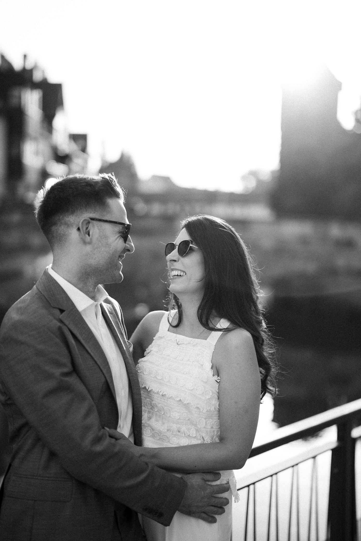 Black and white photo of a smiling couple in sunglasses embracing on a bridge at sunset.
