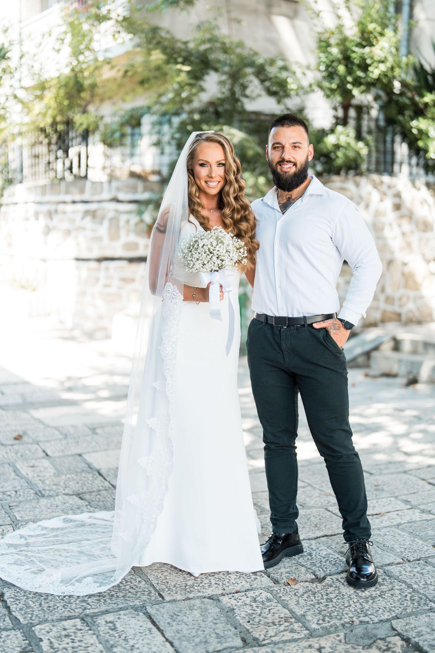 Bride in white gown holding bouquet stands beside bearded groom in dark pants outdoors on stone plaza.
