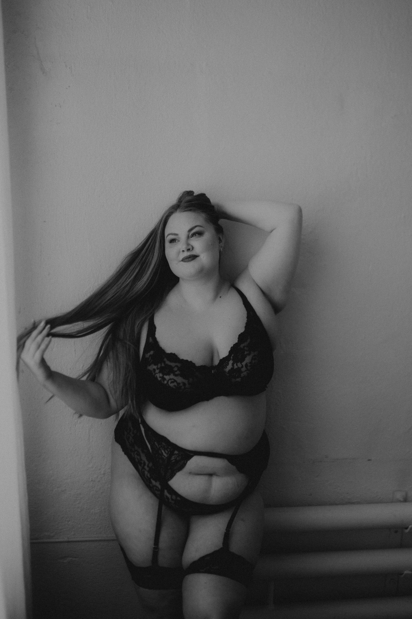 mustavalkoinen boudoir valokuva jossa nainen poseeraa alusvaatteissa seinää vasten.