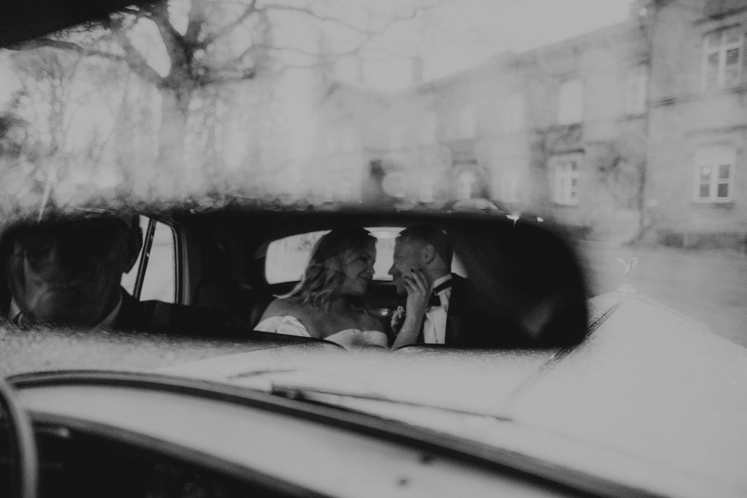 Mustavalkoinen panoraamanäkymä hääparista auton peilistä, jota reunustavat pysäköidyt autot ja rakennukset.