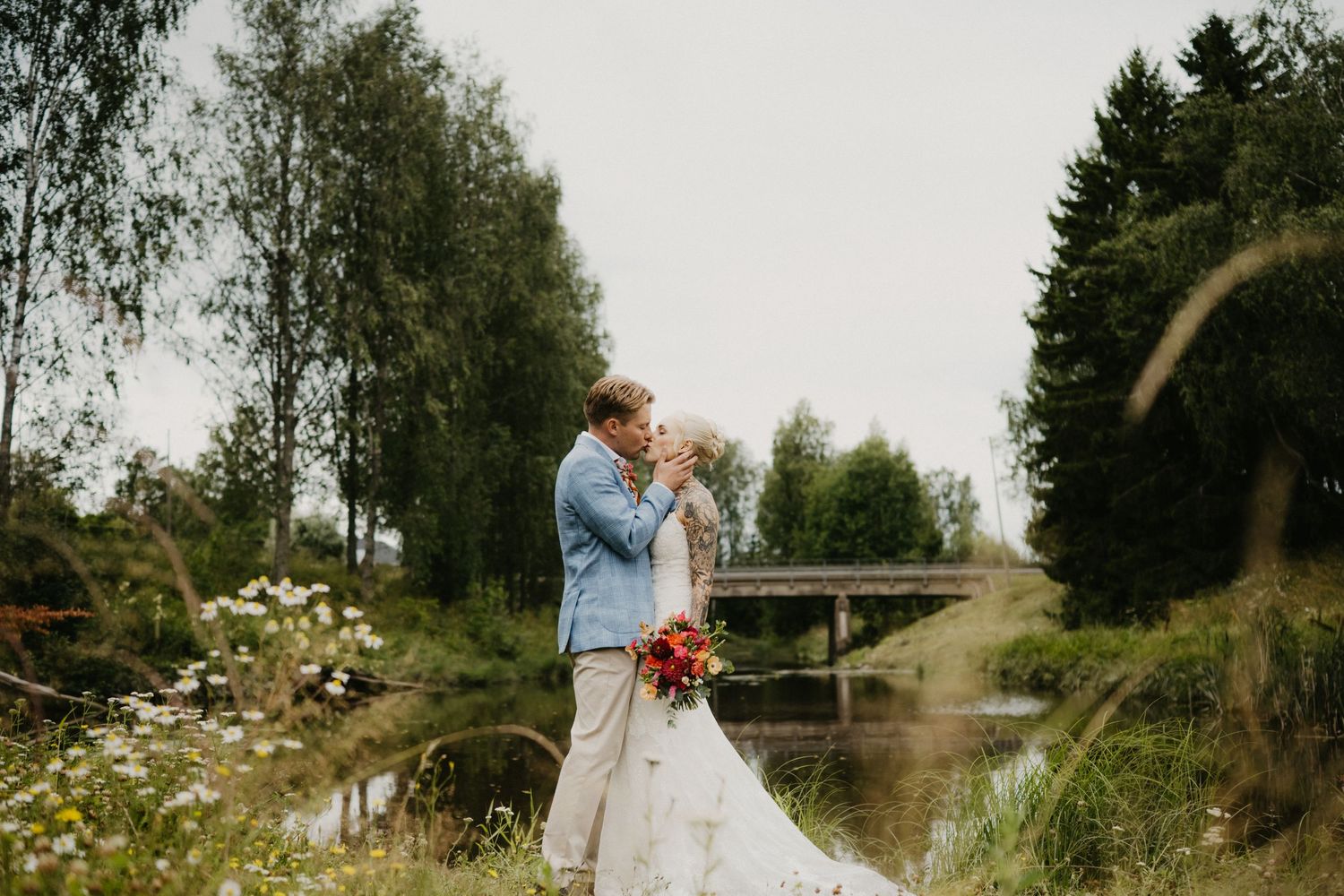 Morsian ja sulhanen vaihtavat romanttisen suudelman ulkona lammen lähellä, luonnonkukkien ja vehreiden puiden ympäröiminä.
