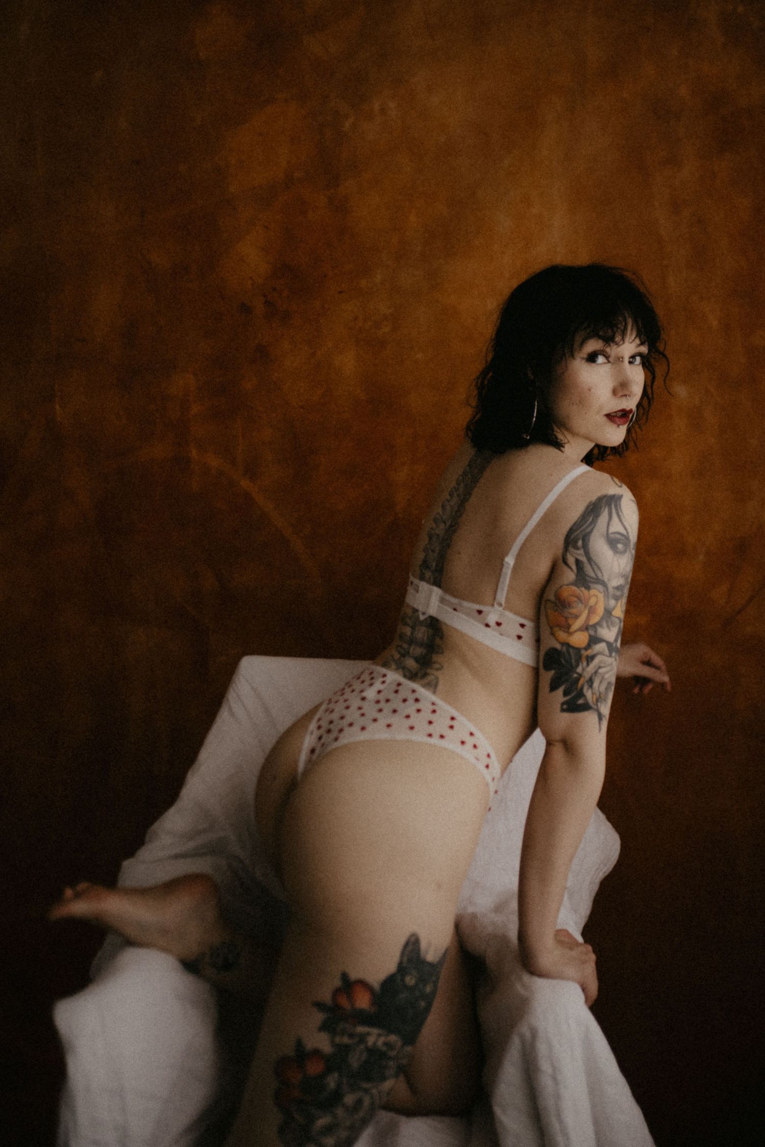 Taiteellinen boudoirkuvas, jossa näkyy hahmo valkoisissa alusvaatteissa, naisella on näkyviä tatuointeja tummanruskeaa taustaa vasten.