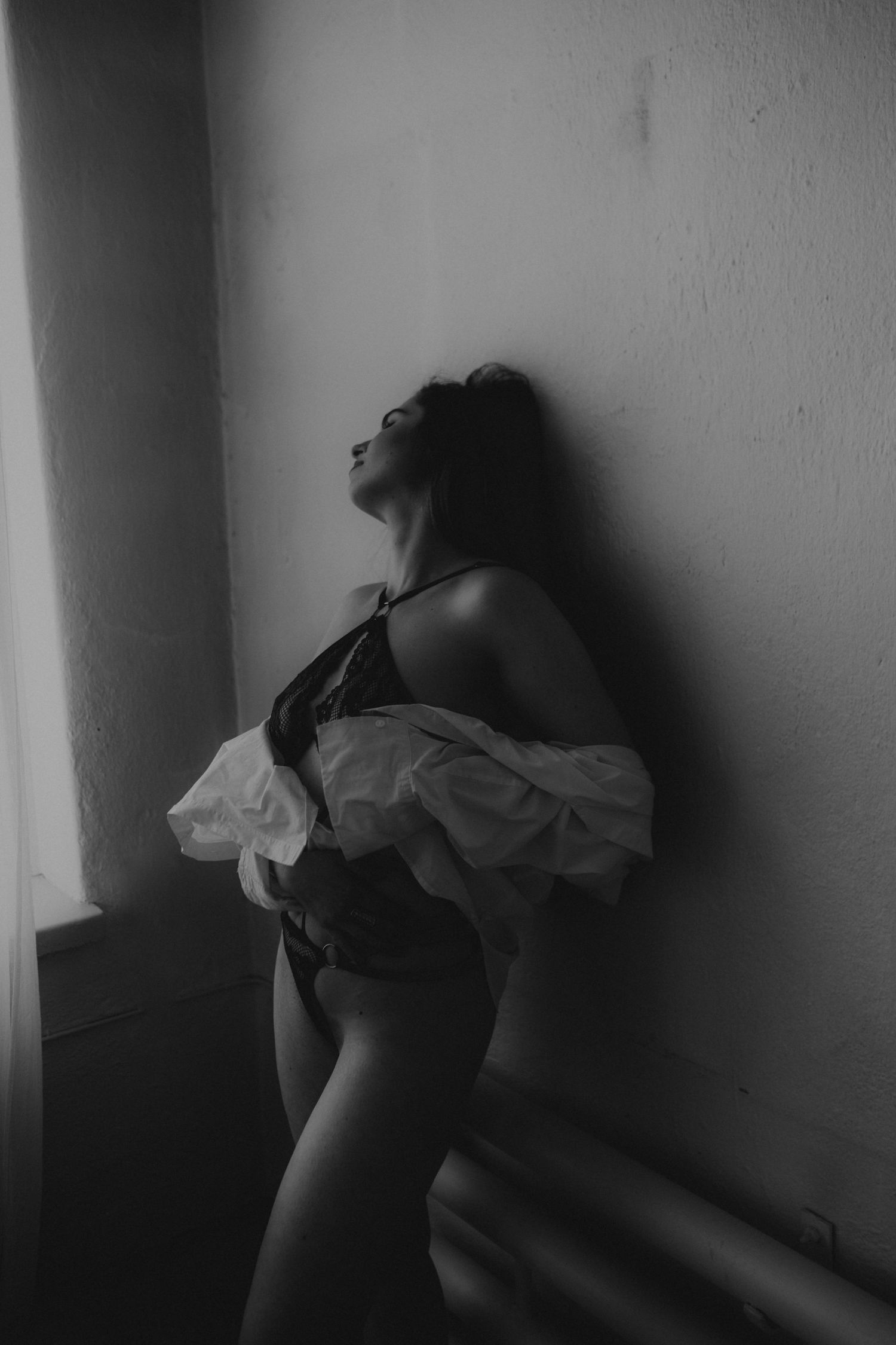 Mustavalkoinen boudoir valokuva, jossa henkilö nojaa seinää vasten seinää tummemmissa varjoissa.