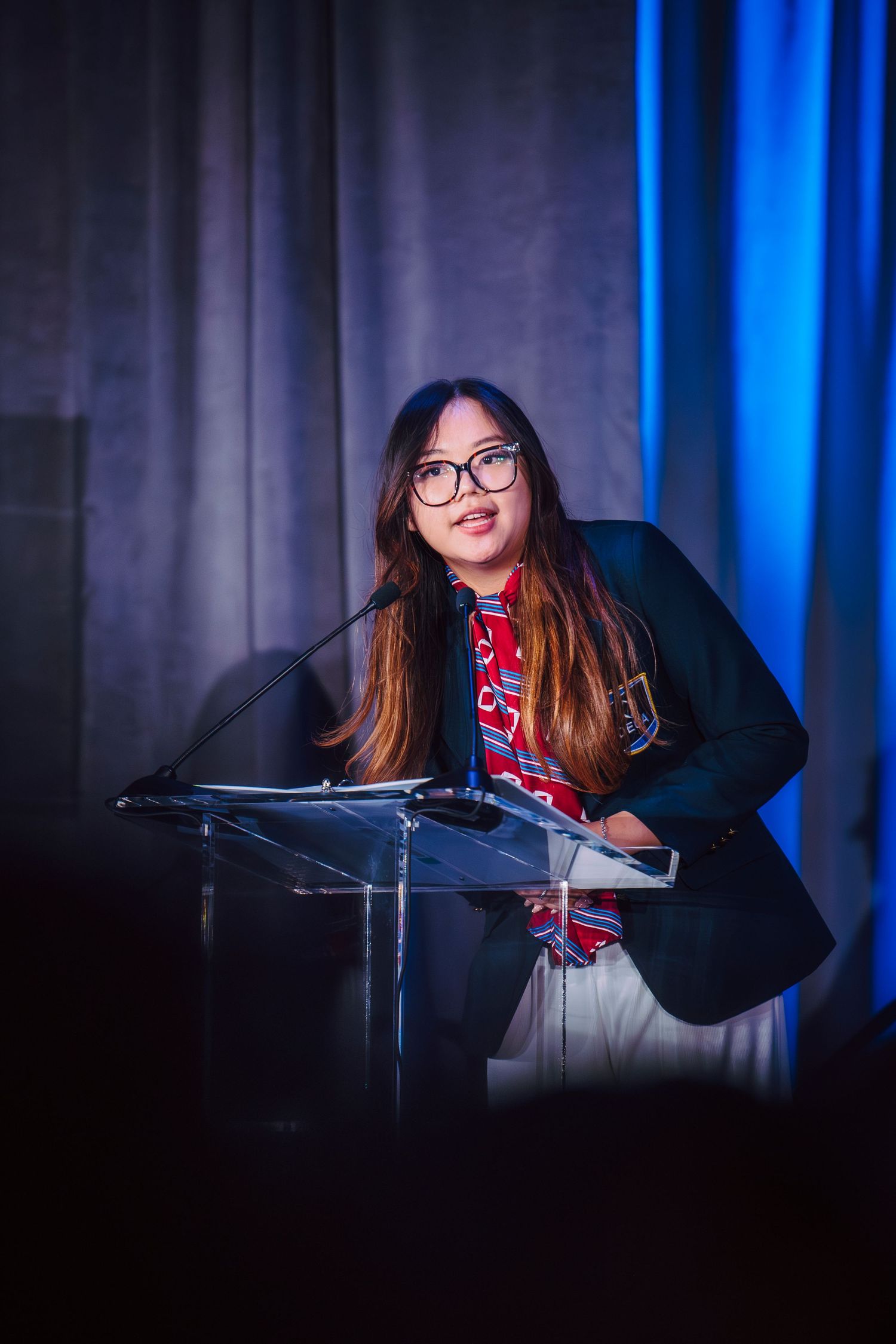Young woman with glasses speaking at a podium during a formal event, wearing a blazer and red scarf.