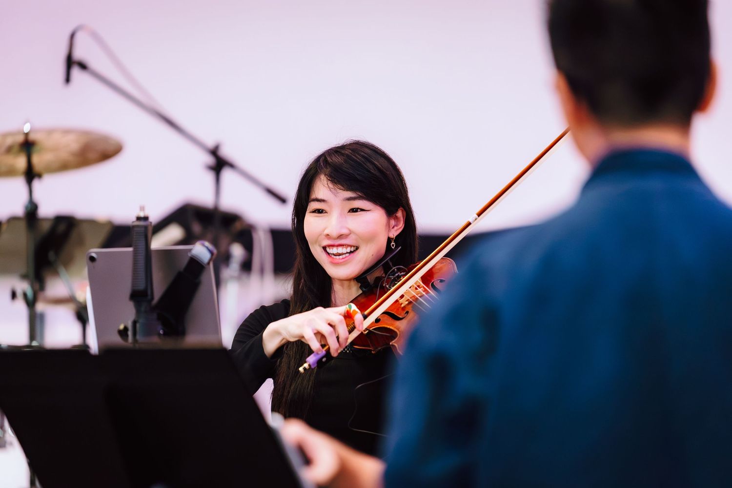 Smiling Asian woman playing violin during a music rehearsal with a drummer and music stand visible in background.