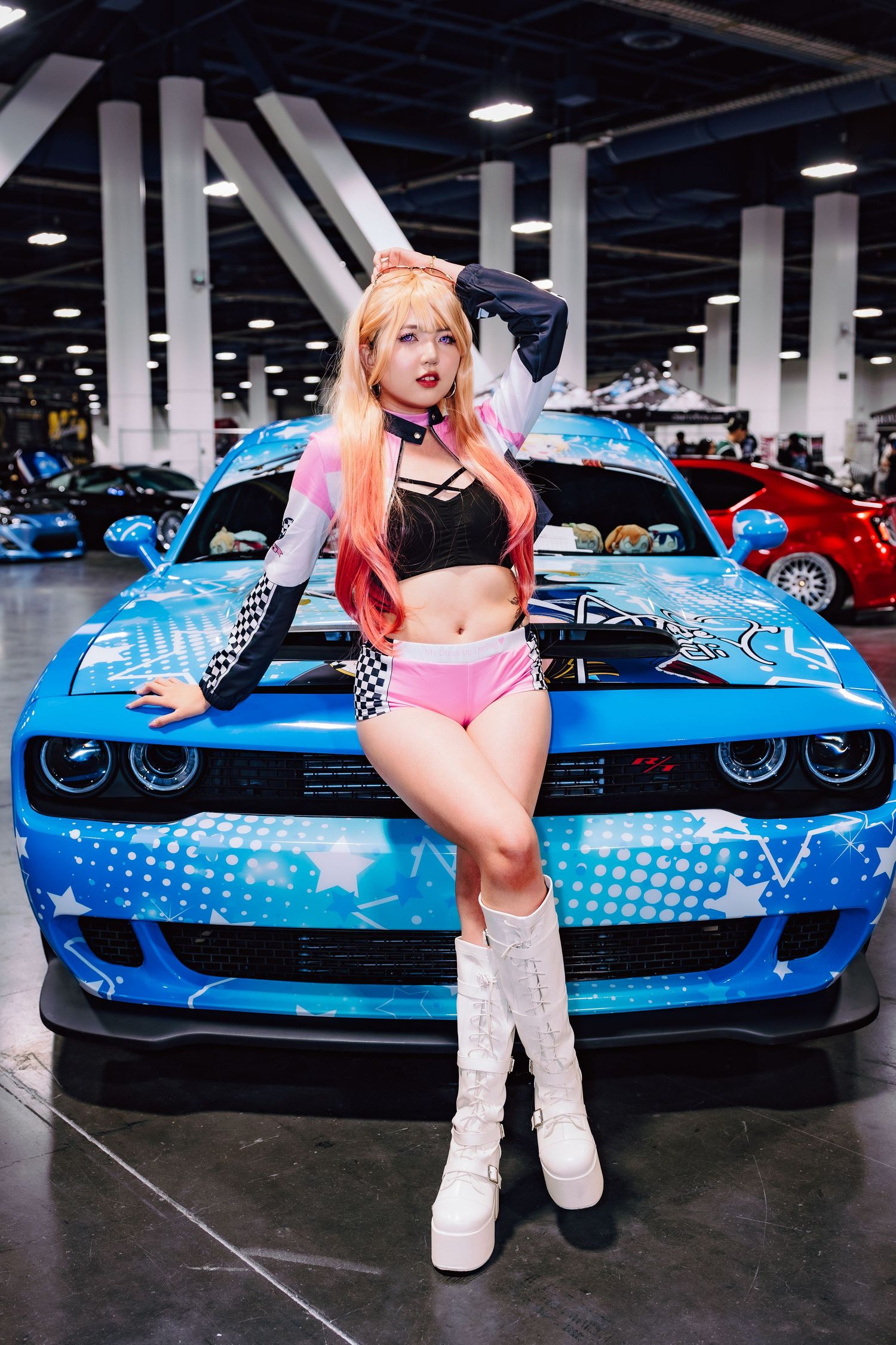Cosplay model in pink wig and crop top posing in front of a blue custom Dodge Challenger at an indoor car show.