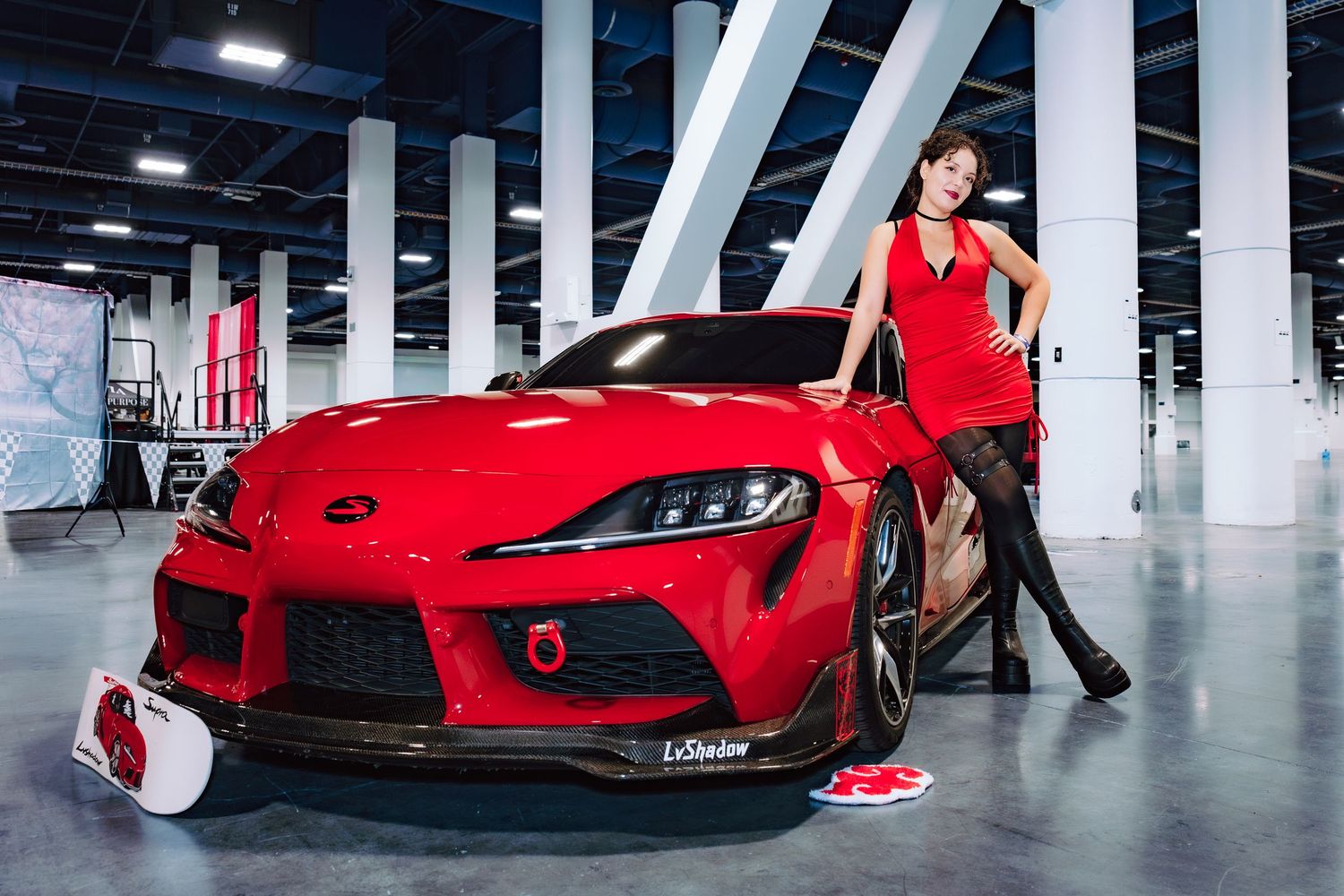 A woman in a red dress poses beside a red Toyota Supra GR at an indoor auto show display.