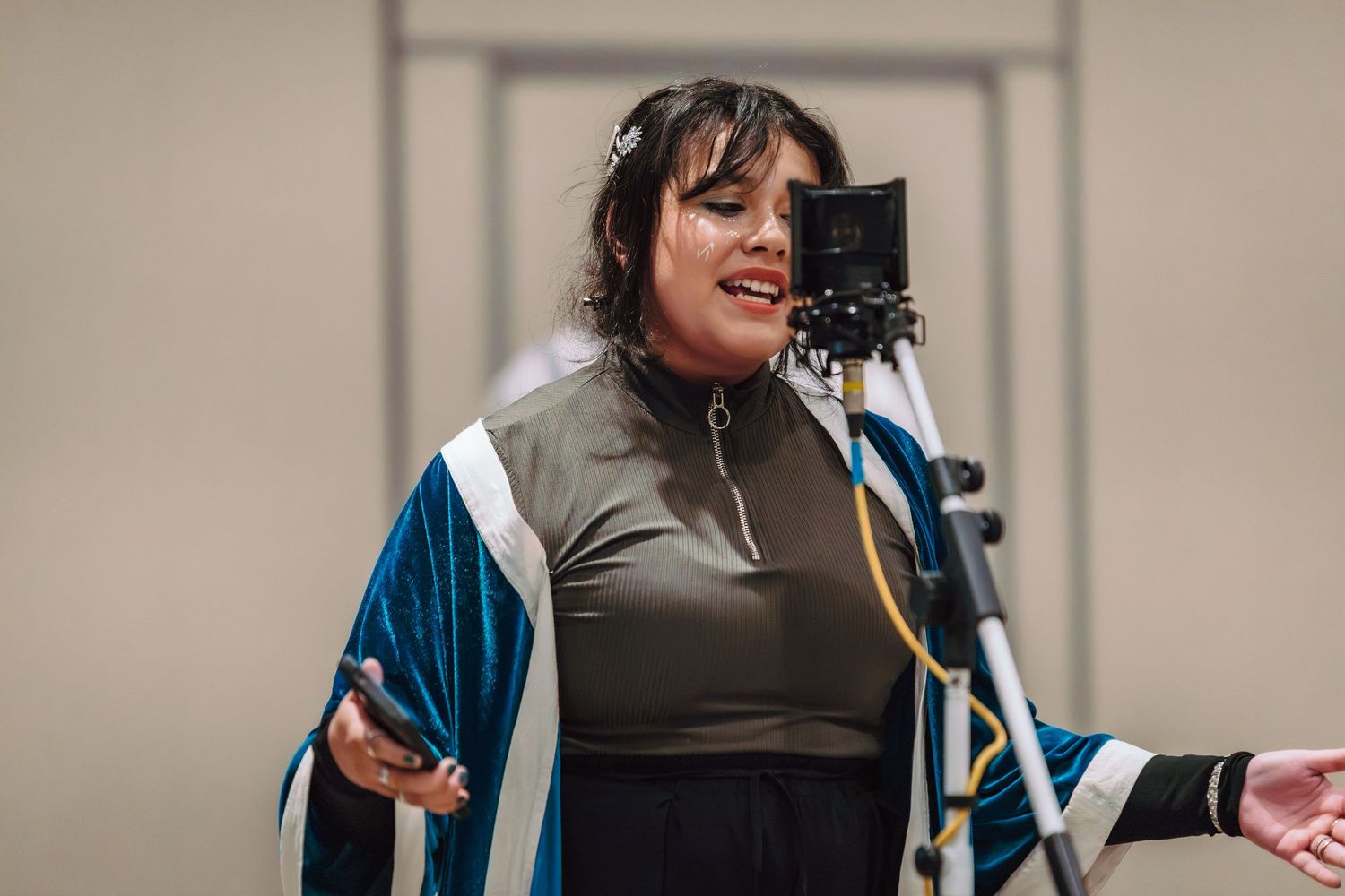 Woman singing into a studio microphone wearing a blue and white robe over a dark outfit.