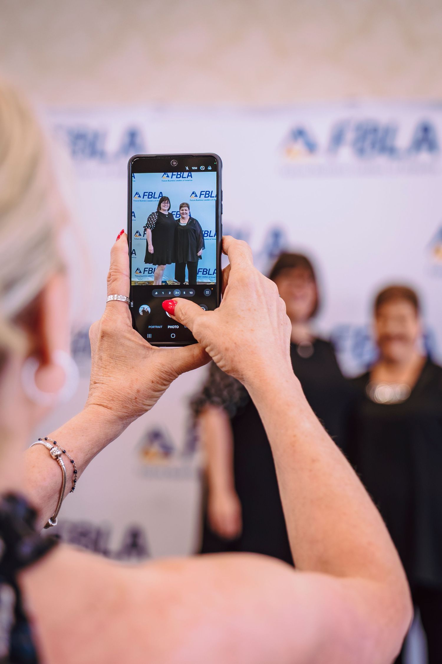 Woman photographs two people posing in front of an FBLA backdrop at a formal event.