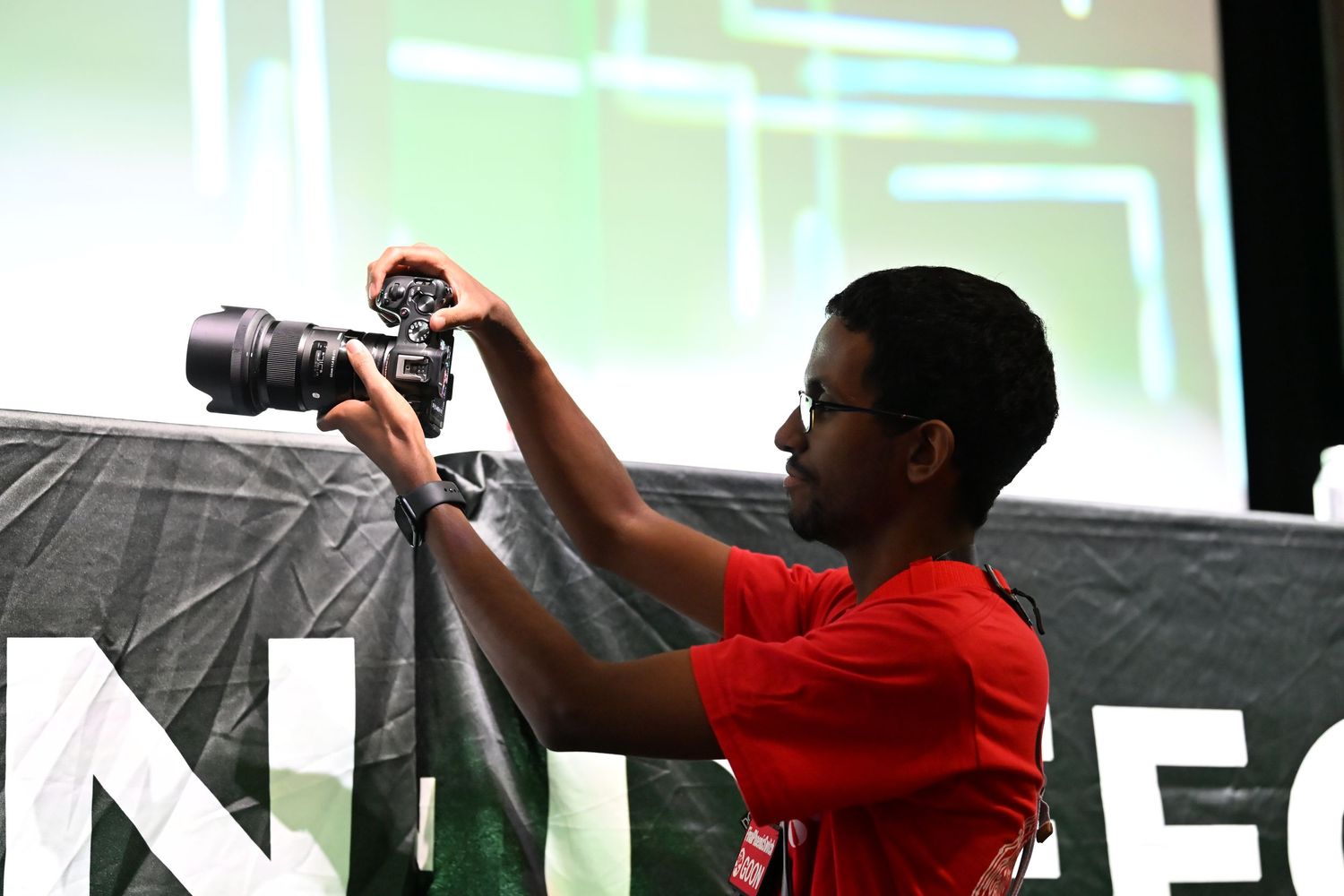 Young photographer in red shirt capturing images with DSLR camera at an event.
