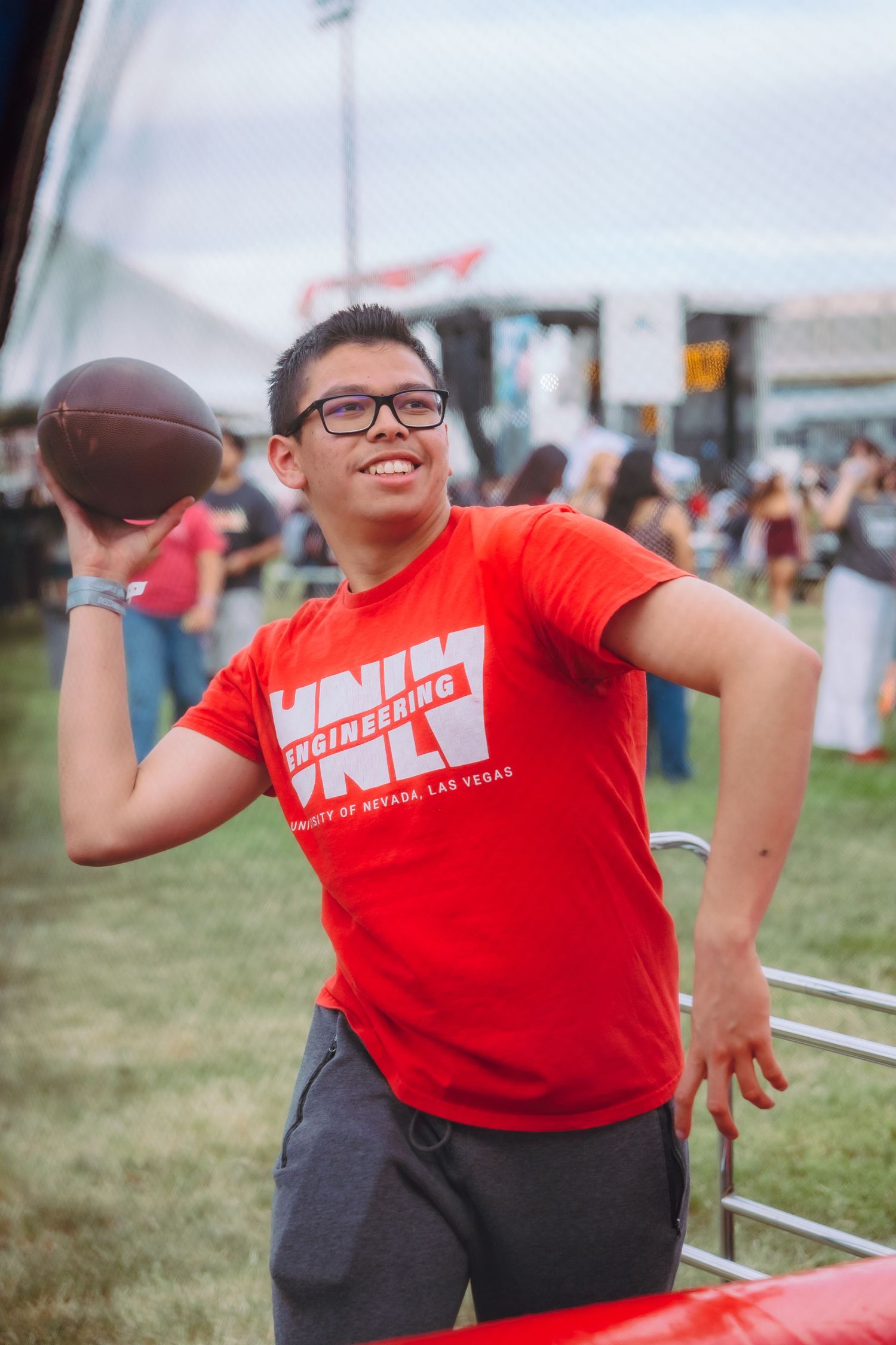 Young man in red UNLV Engineering shirt poses with football at outdoor event on green field.
