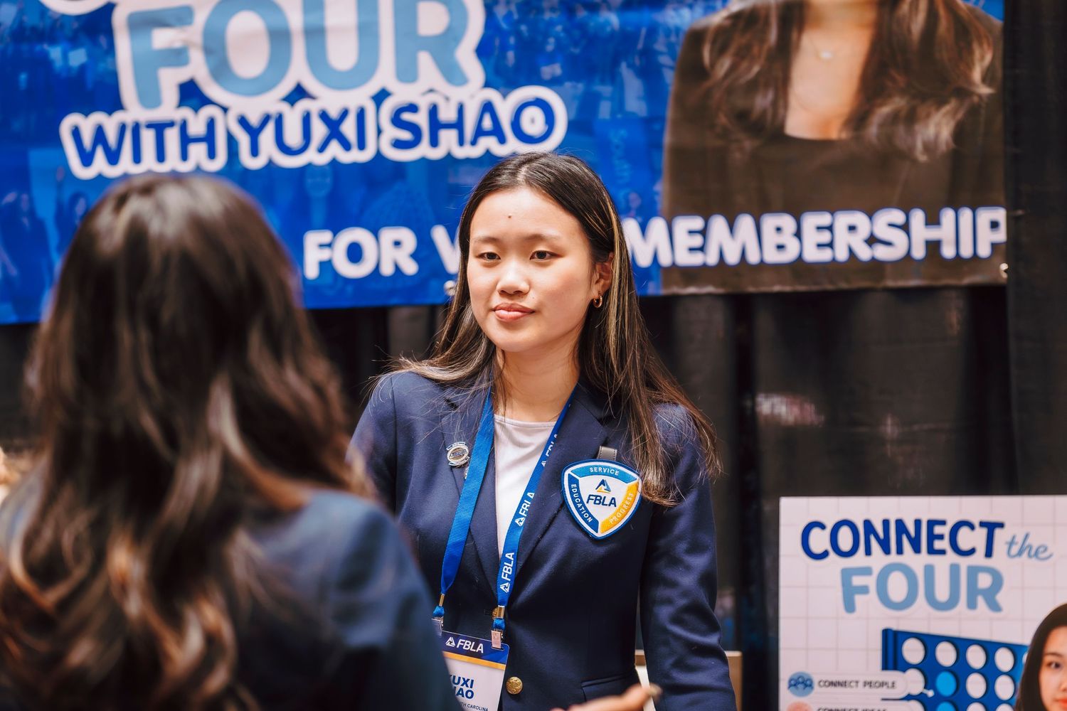 Young woman in FBLA blazer stands at a Connect the Four membership campaign booth at conference.