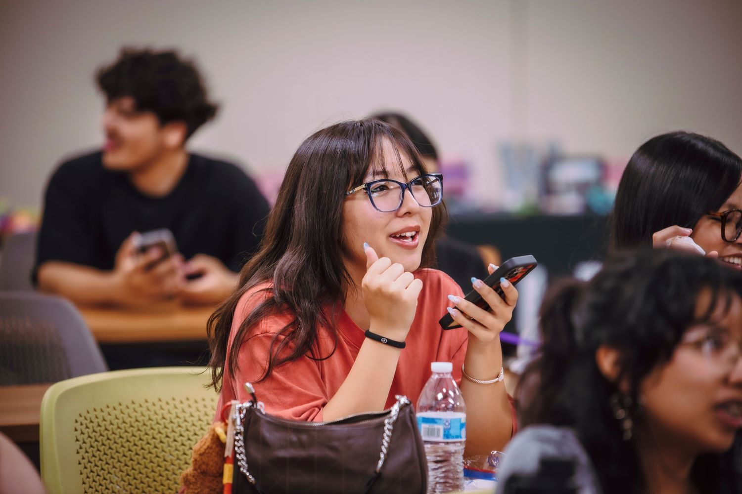 Young woman with glasses smiling and holding a smartphone in a classroom setting with other students.