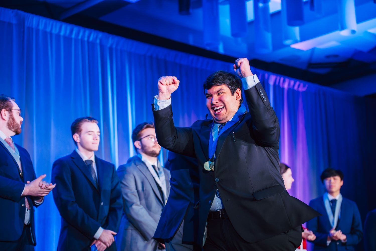 Man celebrating with raised fists at formal event, others in suits applauding on stage with blue lighting.