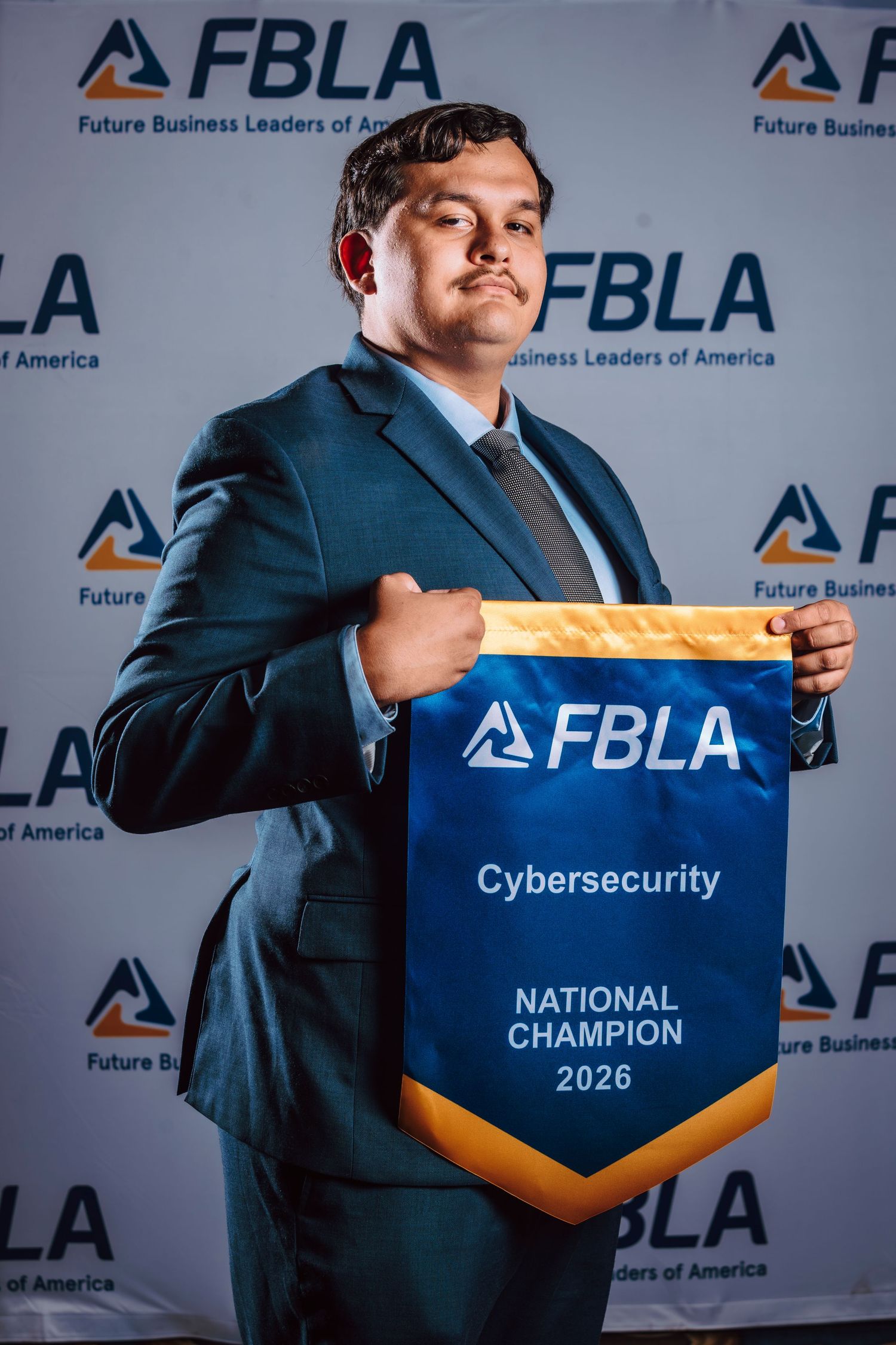 Young man in suit holds FBLA Cybersecurity National Champion 2026 banner in front of FBLA backdrop.