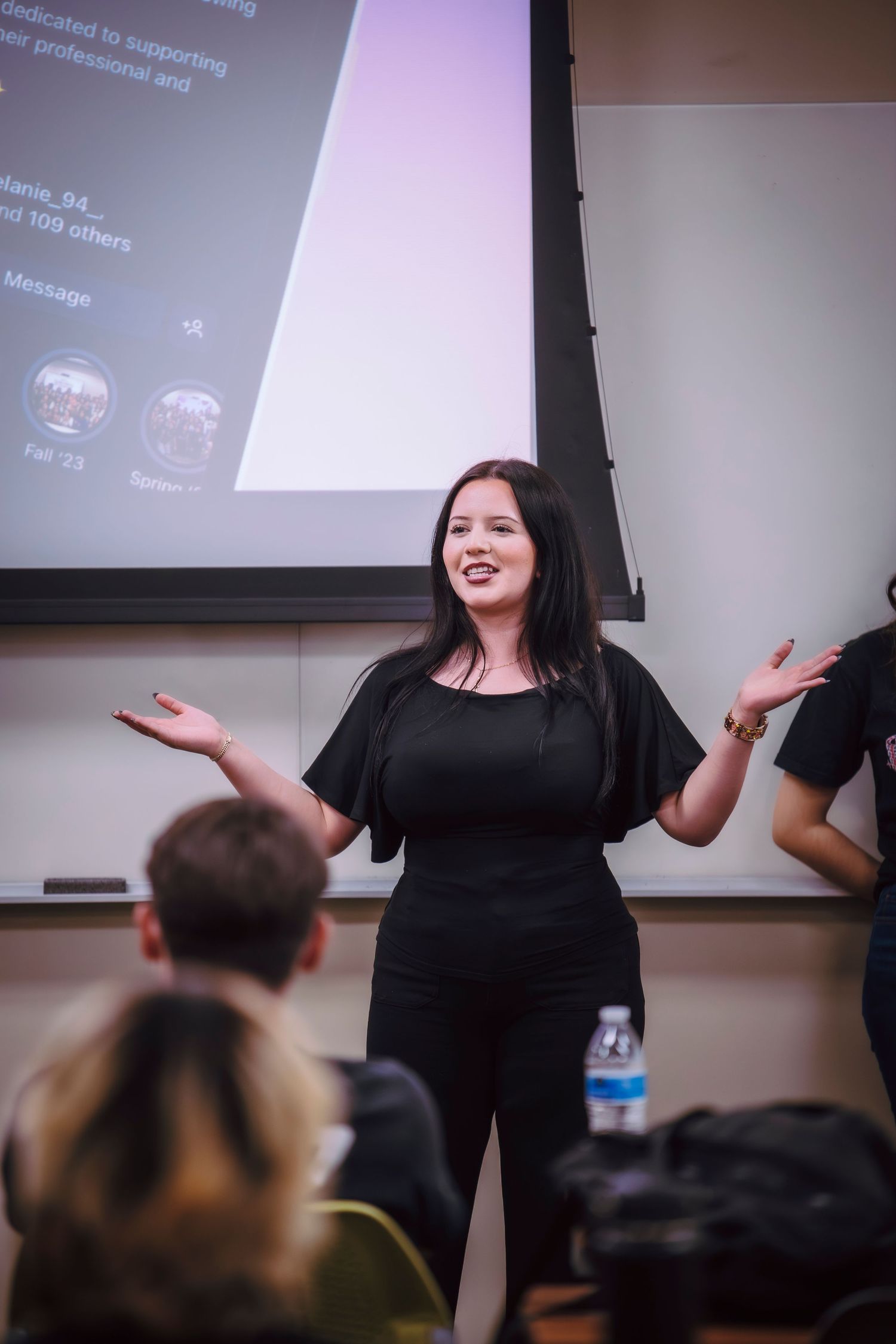 Woman in black outfit presenting to an audience in a classroom with a projected social media screen behind her.