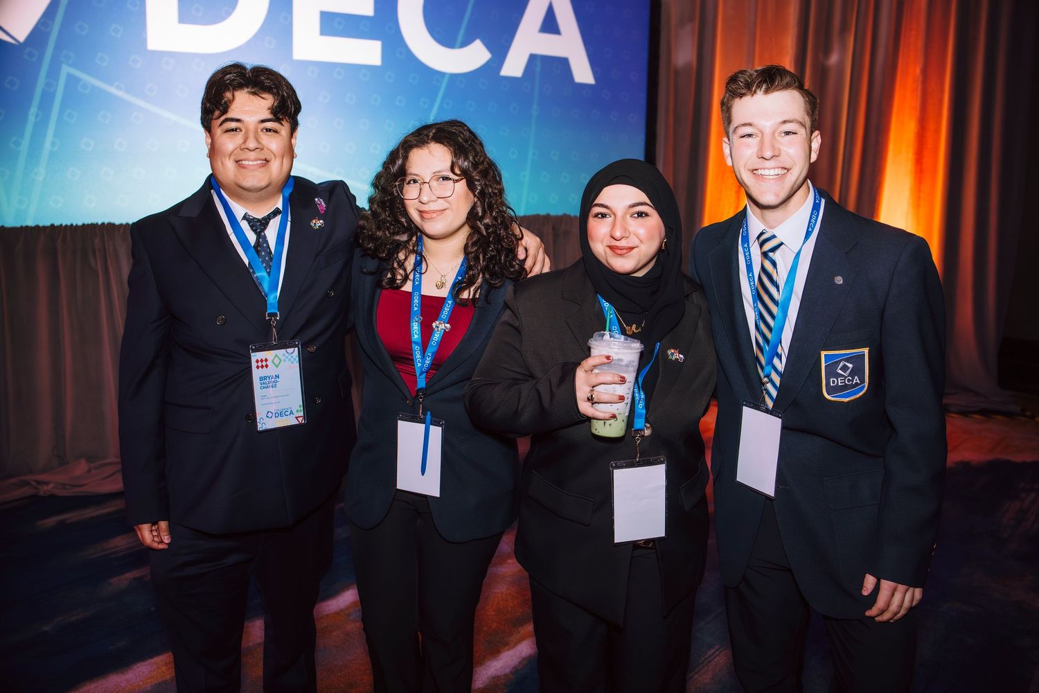 Four DECA students in business attire smile together at a DECA conference event with blue backdrop.