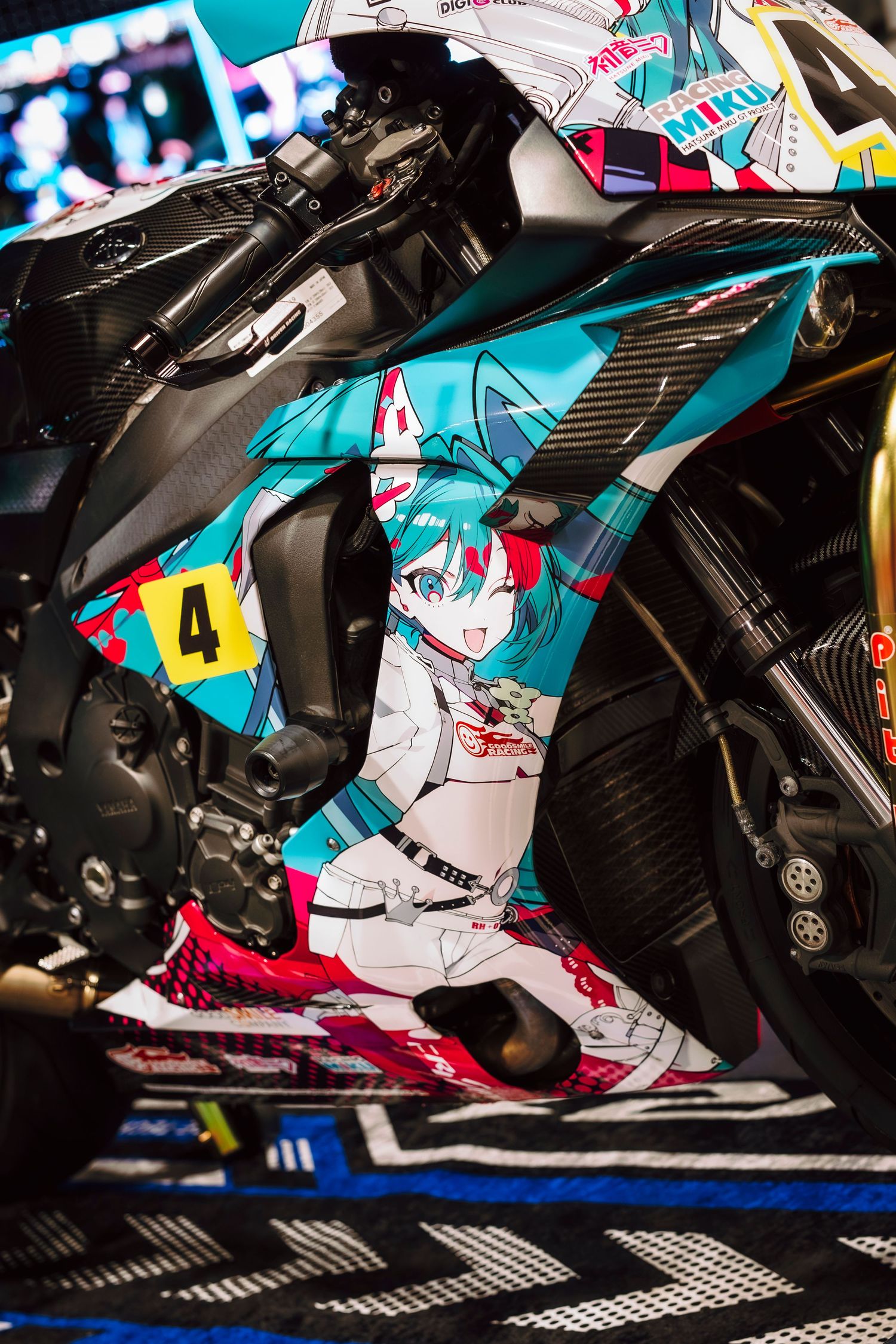 Racing motorcycle with Hatsune Miku anime artwork wrap, featuring teal hair character design and number 4, displayed at motorsport event.