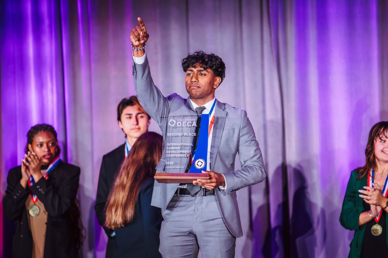 Young man raises fist holding award plaque on stage with purple lighting at academic achievement ceremony.