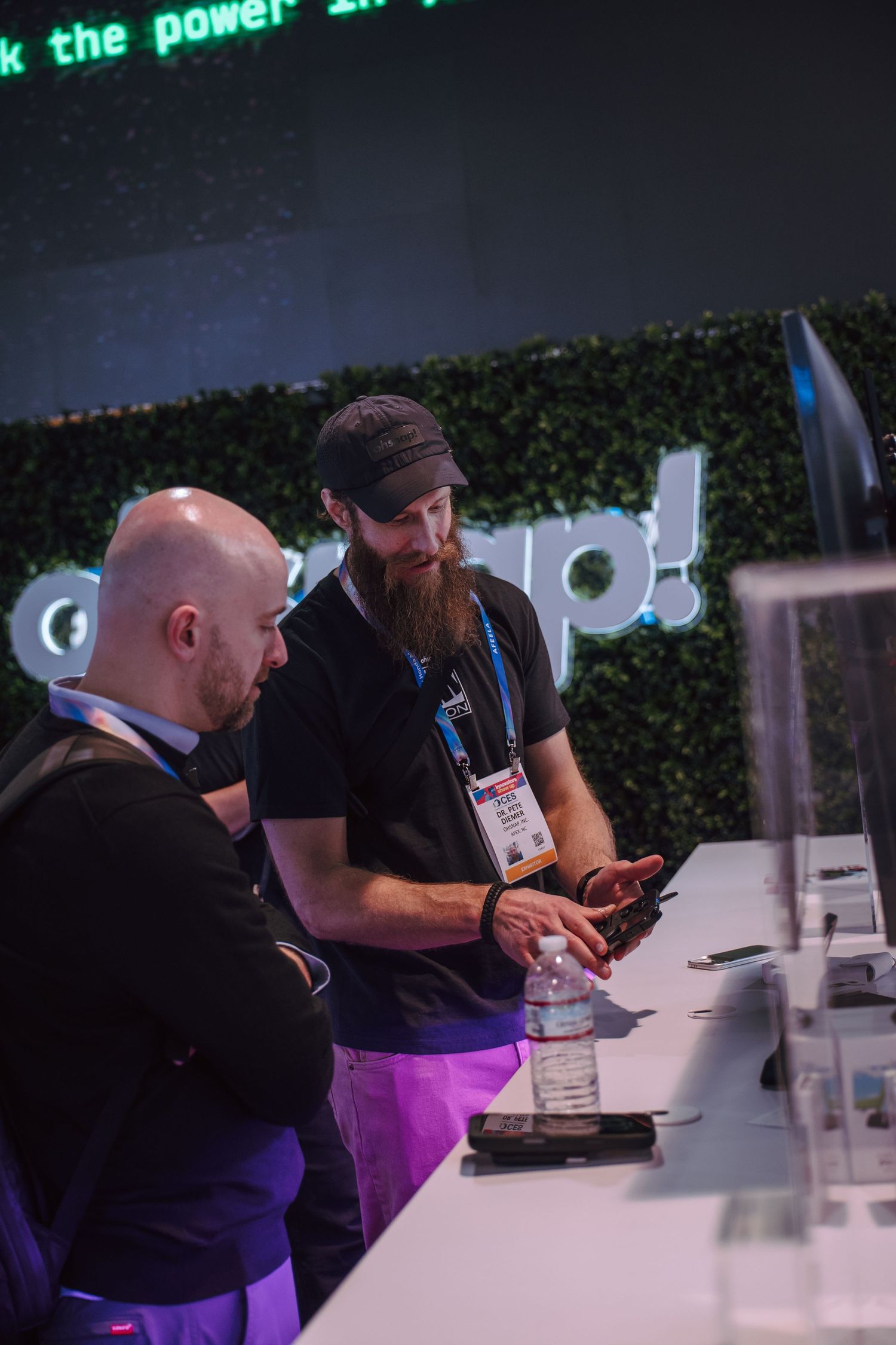 Two men at a tech expo booth examine a device, with a green living wall and neon signage in the background.