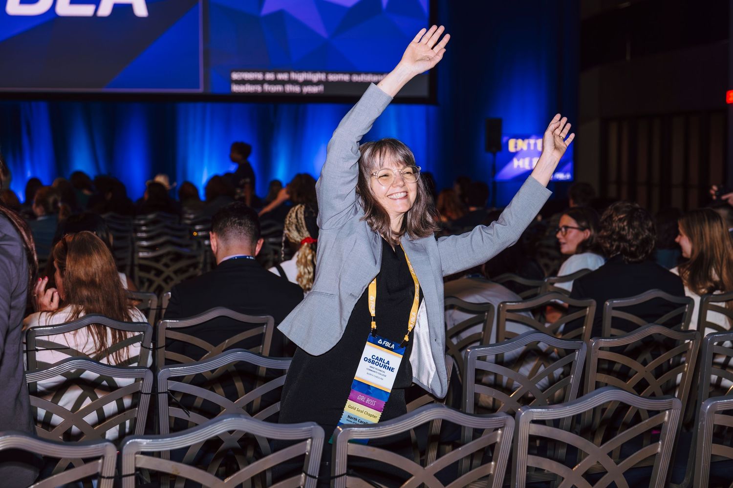 Woman with arms raised celebrating at a professional conference event with blue stage lighting.