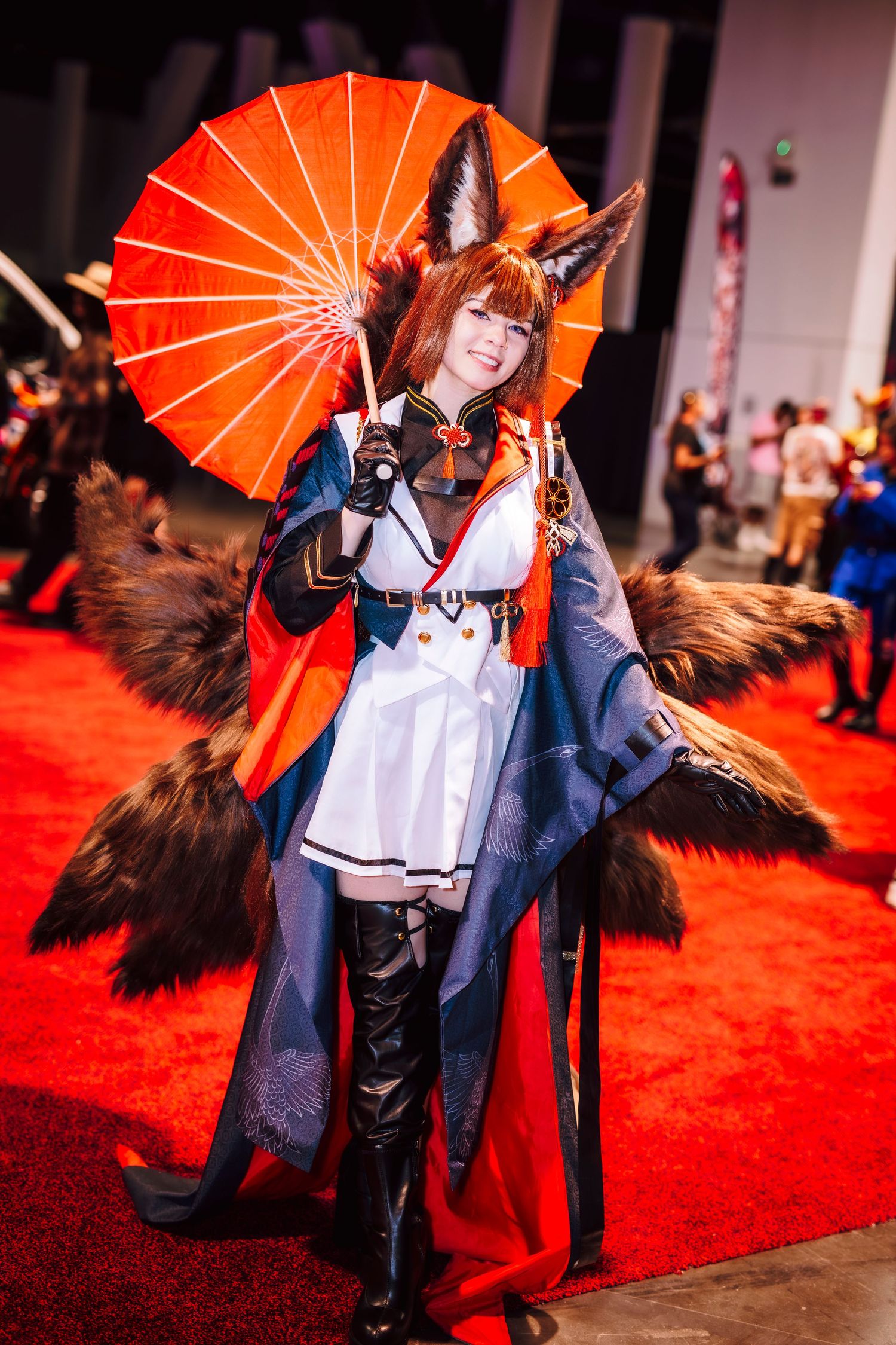 Cosplayer in fox ear costume with fluffy tails, white dress, blue cape, black boots, holding red parasol at anime convention.