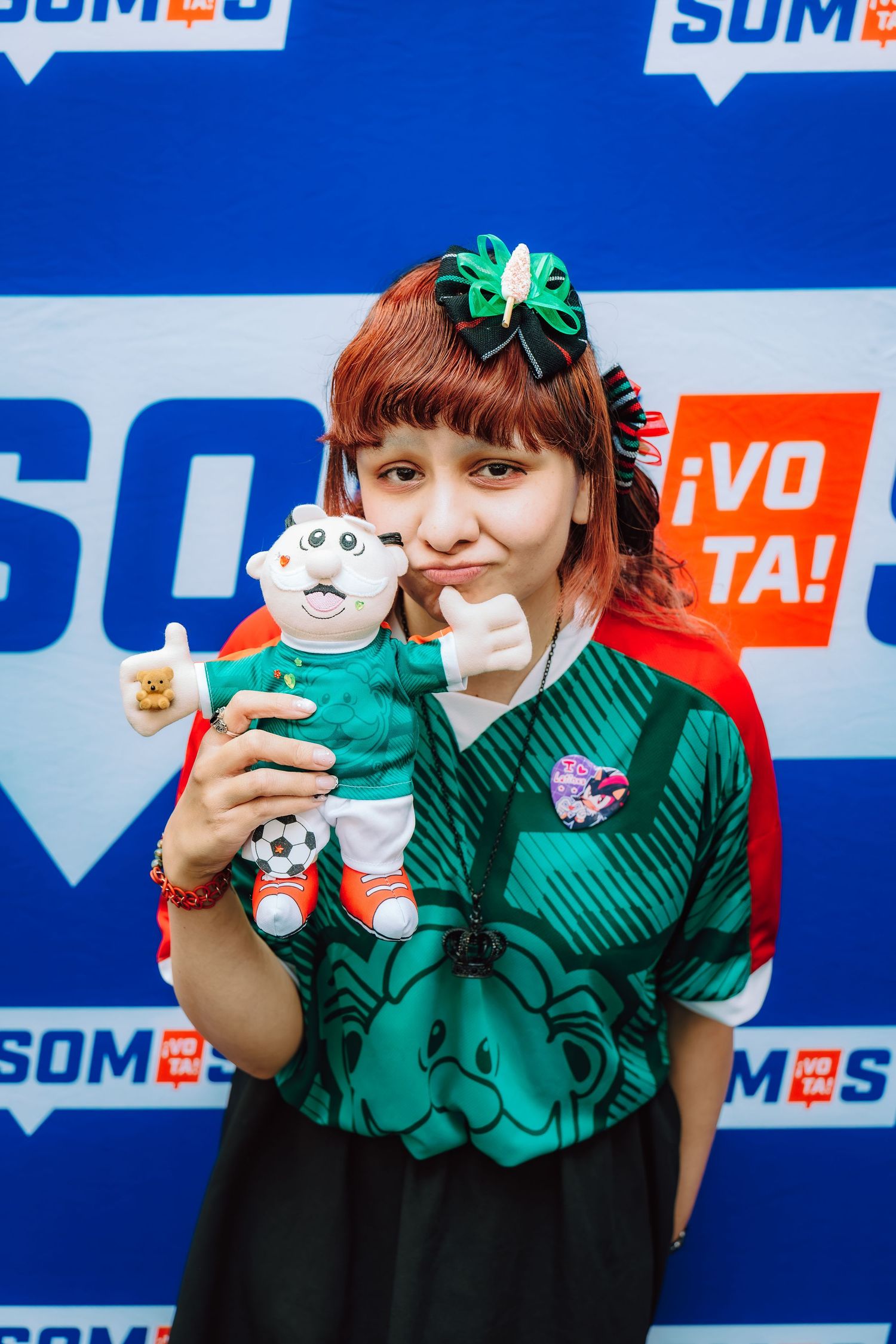 Young woman with red hair holding a soccer mascot plush toy, wearing a green jersey at a Somos Vota event.