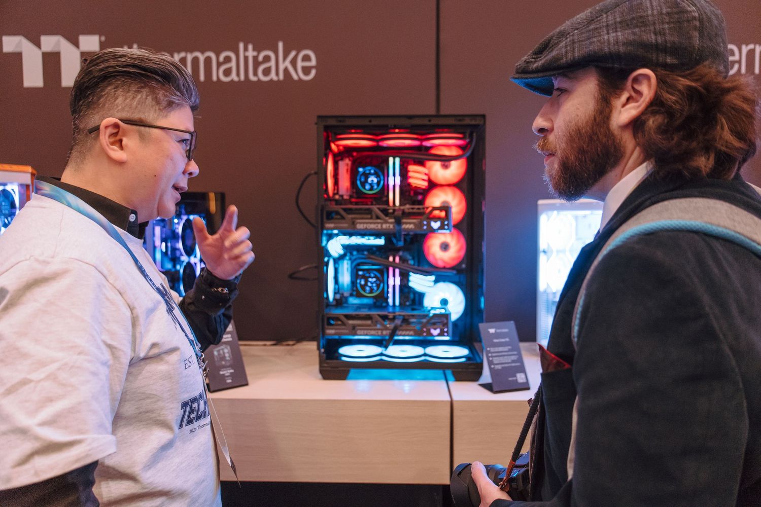Two men discuss a illuminated Thermaltake PC case with red and blue RGB lighting at a tech trade show.