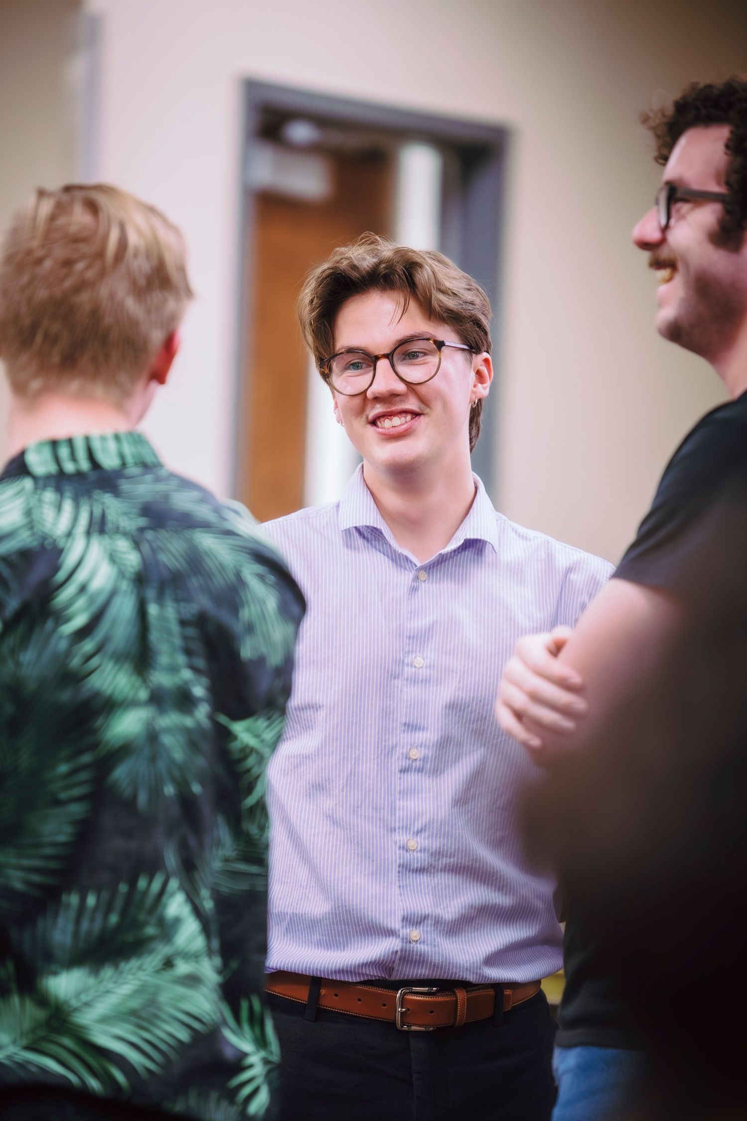 Young man with glasses smiling during a casual conversation at an indoor social gathering.