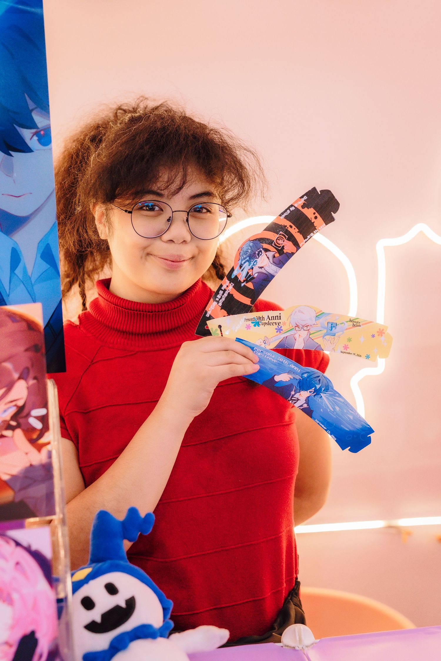 Young woman in red turtleneck and glasses holding anime-themed toy gun at a gaming convention booth.